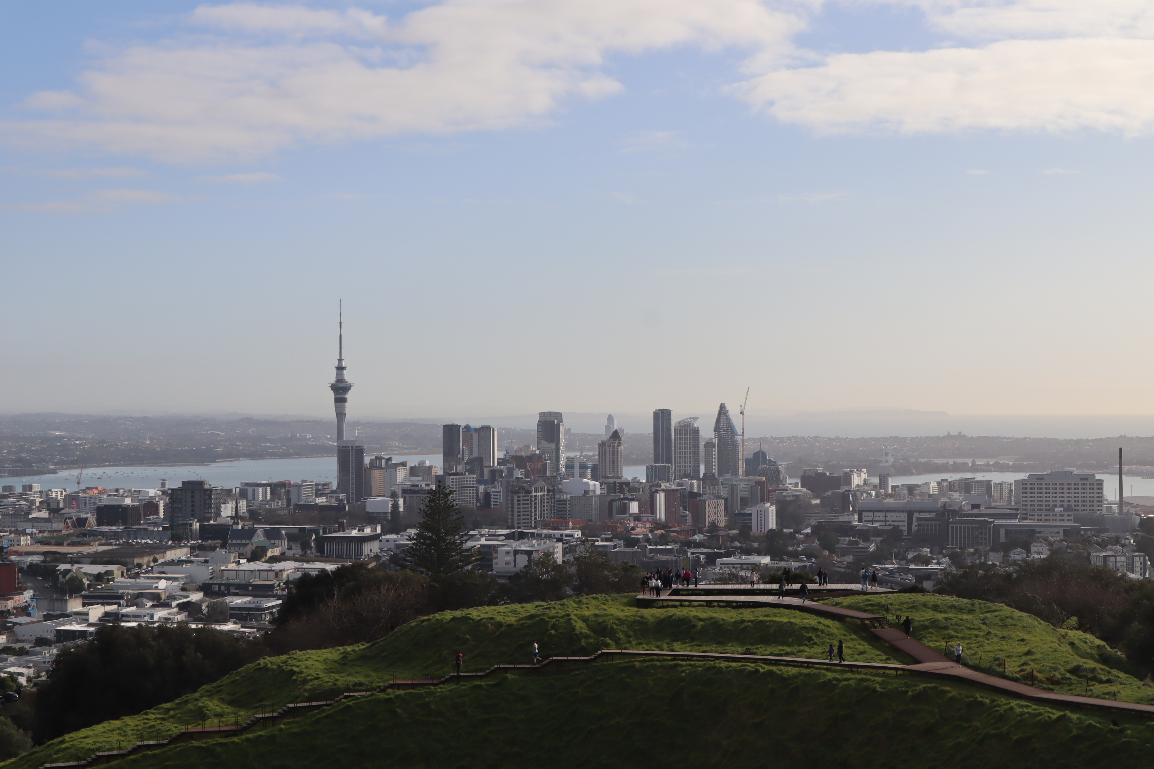 Skyline of Auckland, New Zealand, viewed from a green hill with people walking along a path on a cloudy day