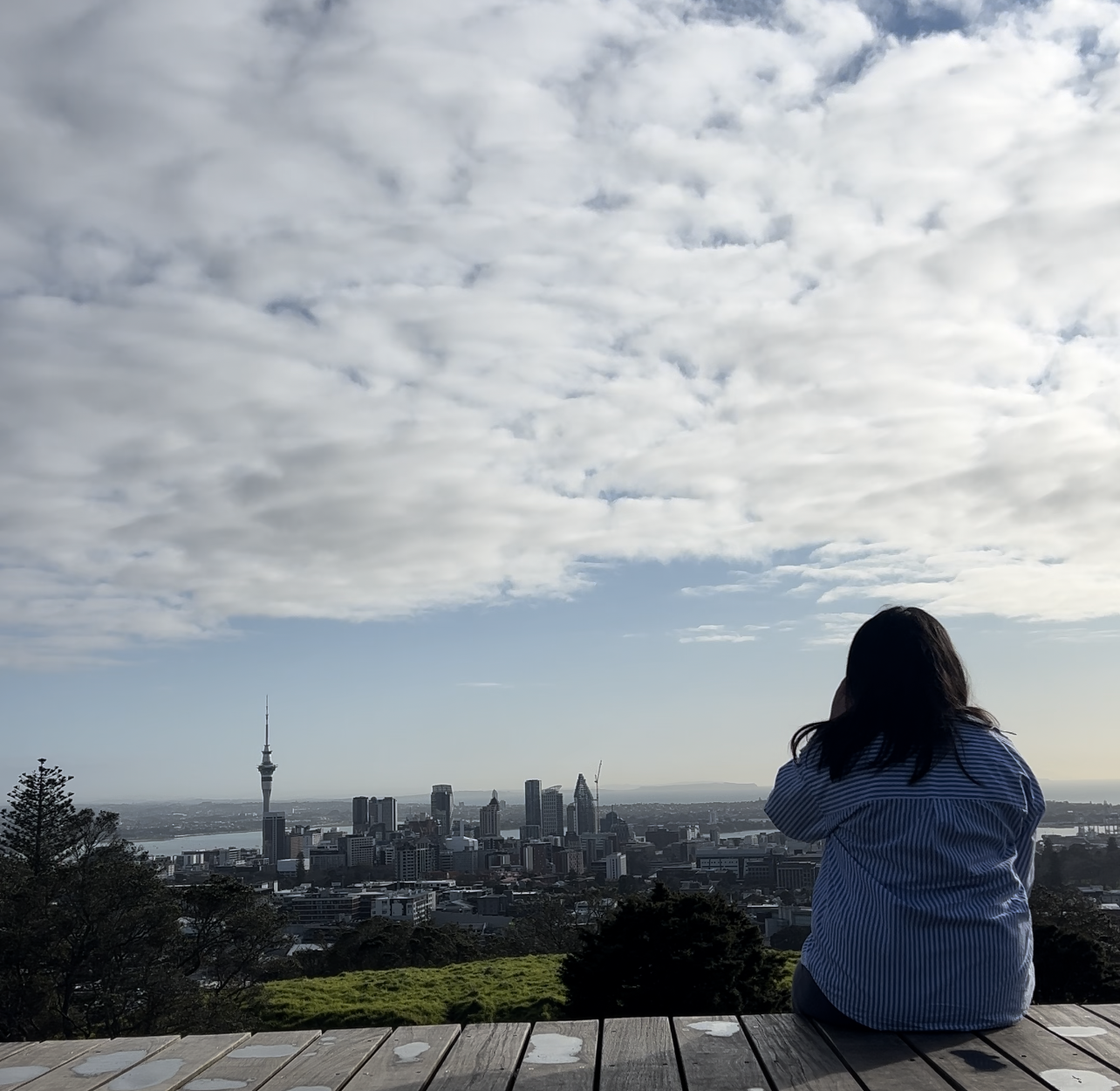 Person sitting on a wooden platform, overlooking a city skyline with Skytower in the distance. The sky is partly cloudy