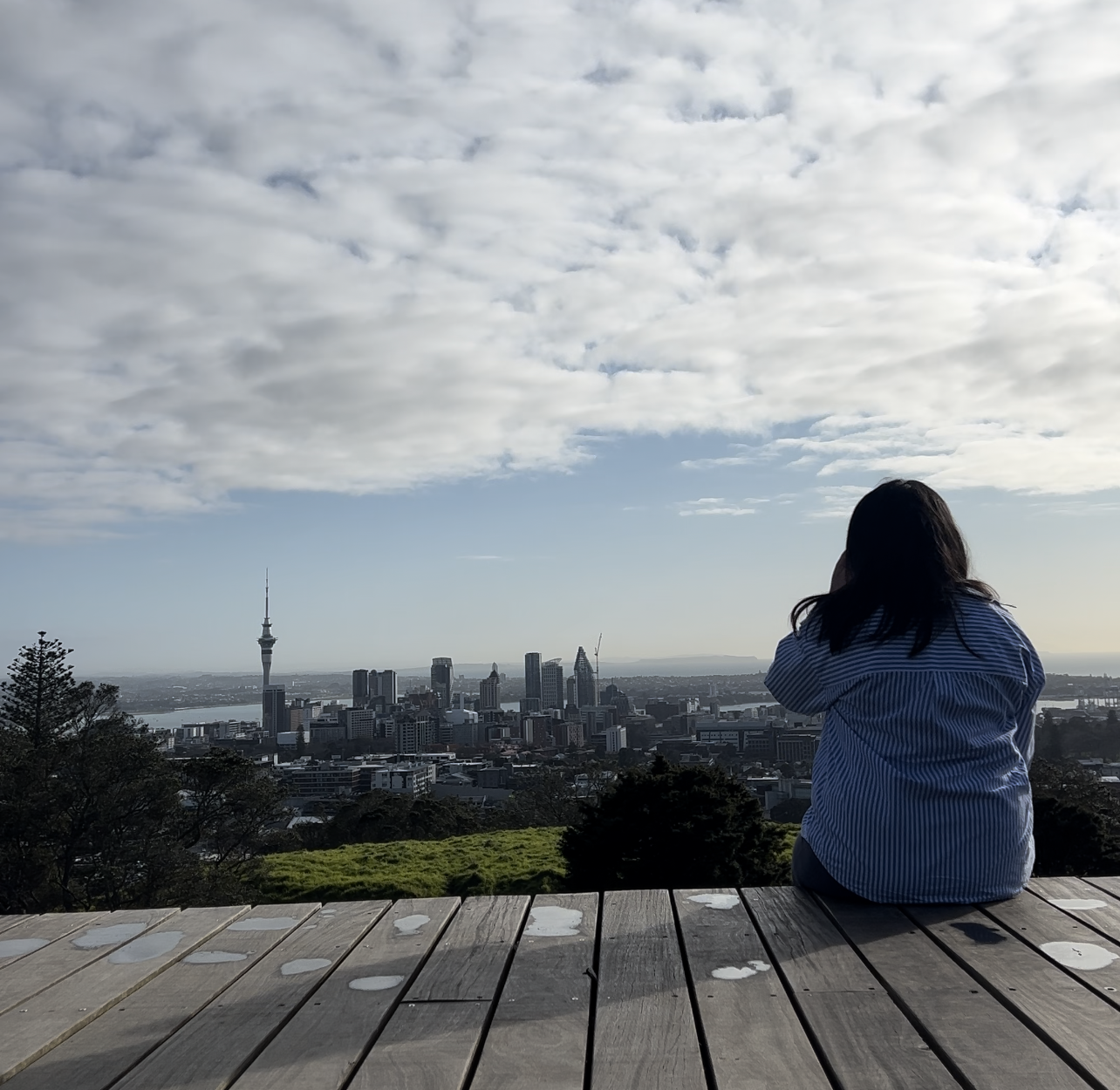 solo trip to japan itinerary Person sitting on a wooden platform, overlooking a city skyline with Skytower in the distance. The sky is partly cloudy