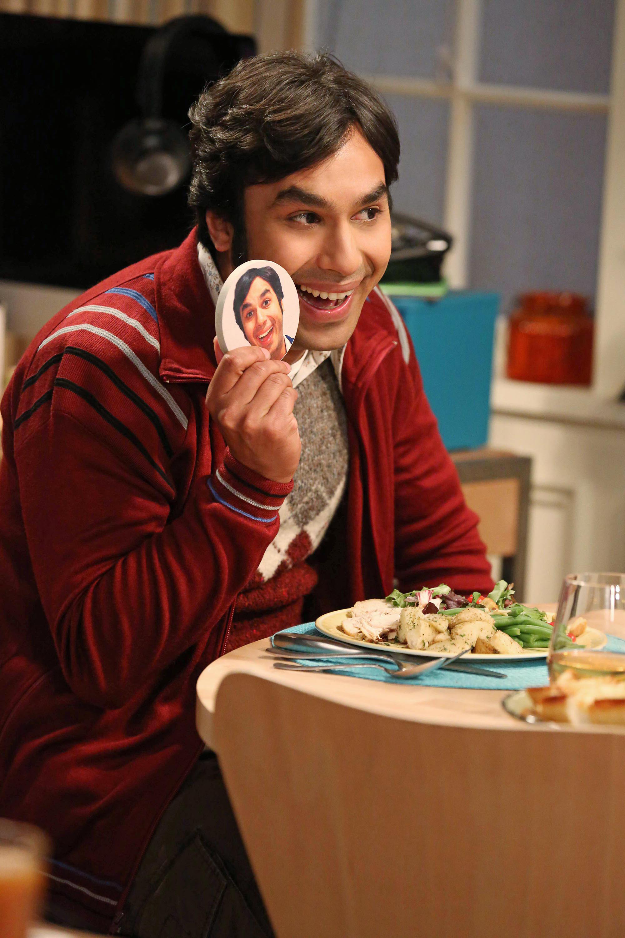 Kunal Nayyar smiles while holding a circular photo of himself at a dinner table with a meal in front of him