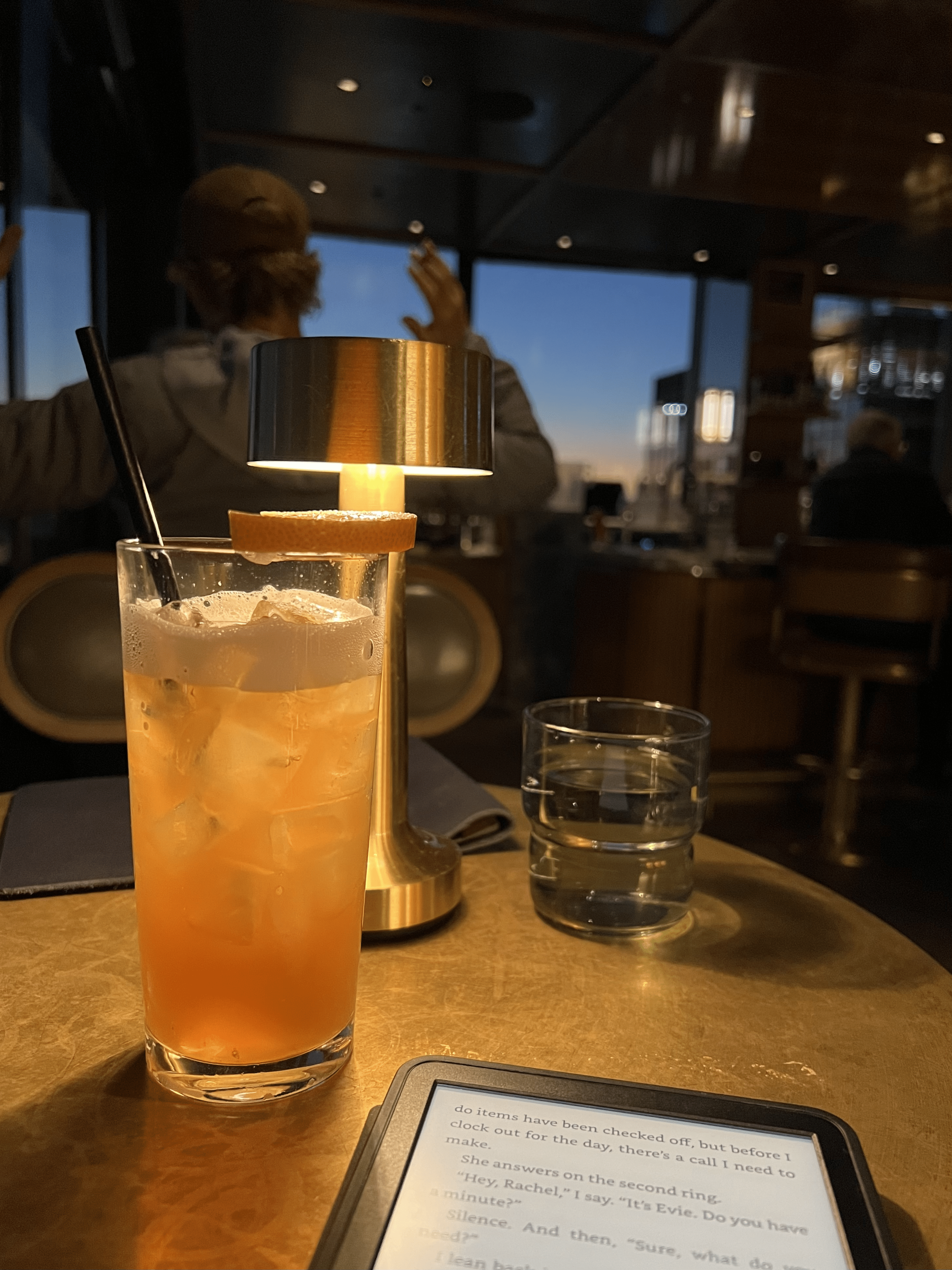 solo trip to japan itinerary A dimly lit bar scene with a person blurred in the background. A cocktail and a glass of water are on the table alongside an e-reader displaying text