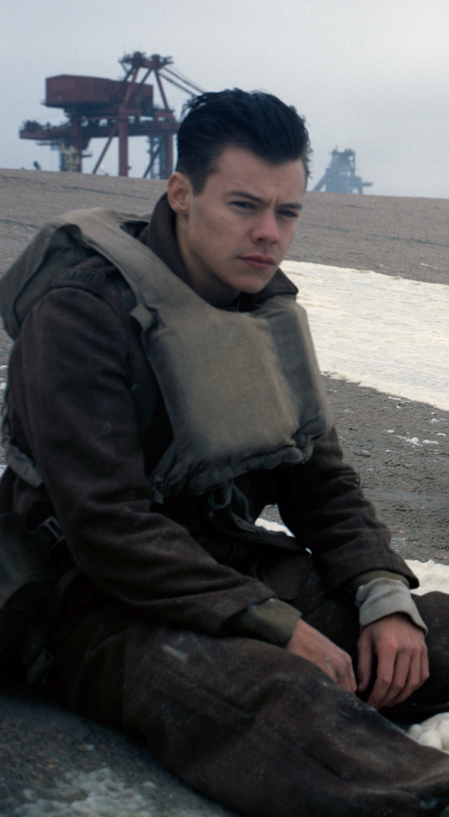 Harry Styles sits on a beach in a military uniform and lifejacket, appearing contemplative. Industrial structures are seen faintly in the background