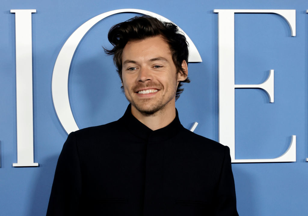 Harry Styles smiles while wearing a dark, tailored suit in front of a blue backdrop with white text at a public event