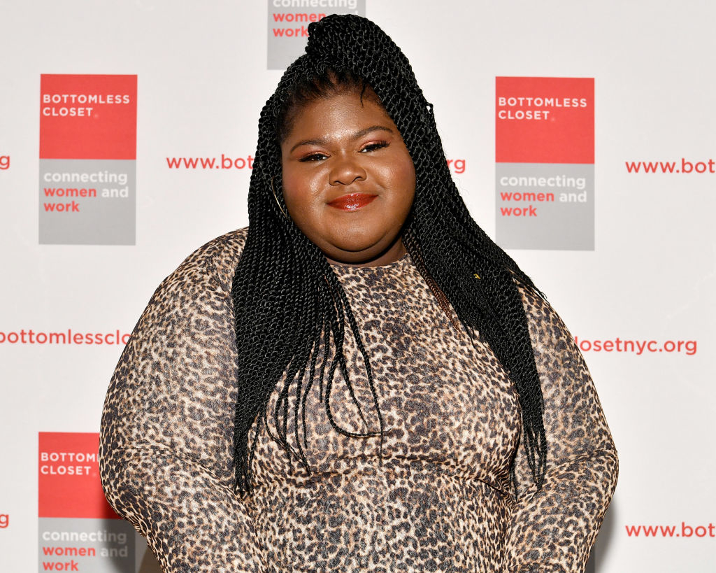 Gabourey Sidibe at a Bottomless Closet event, wearing a leopard print dress and smiling. Bottomless Closet banners in the background