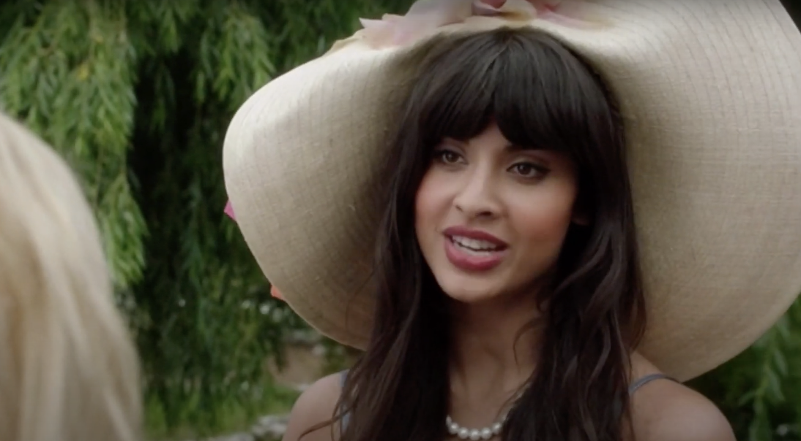 Jameela Jamil wearing a large hat with a floral accent and a pearl necklace, smiling while speaking to a person with visible blonde hair
