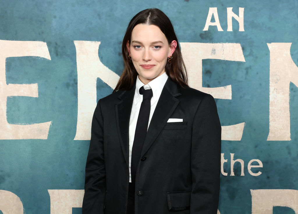Victoria Pedretti in a black suit and tie, stands in front of a promotional backdrop for "A Friend of the Family"
