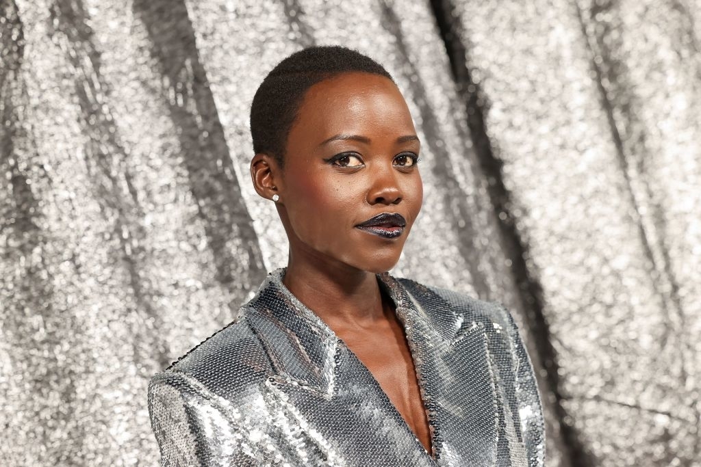 Lupita Nyong'o poses in a sequin-studded jacket, looking at the camera with a confident expression against a sparkling backdrop