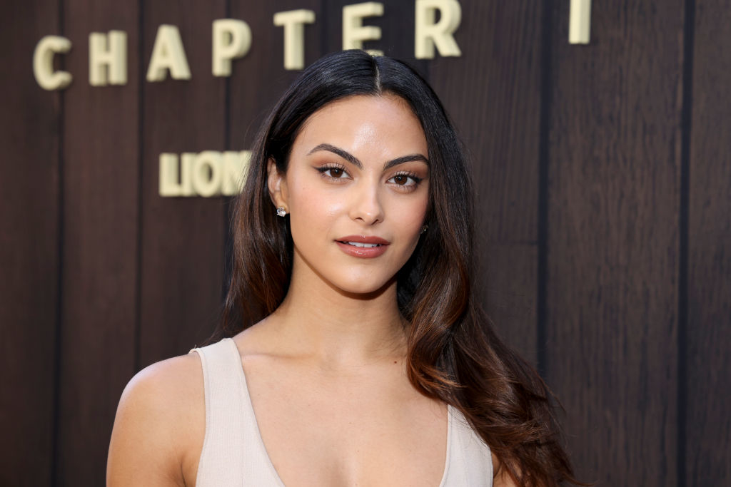 Camila Mendes at a media event, wearing a sleeveless, low-cut top. The background displays text "Chapter" and "Lion"
