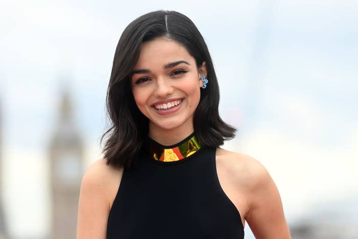 Rachel Zegler is smiling, wearing a sleeveless black top with a gold collar detail, and blue earrings. She has shoulder-length hair and is standing outdoors