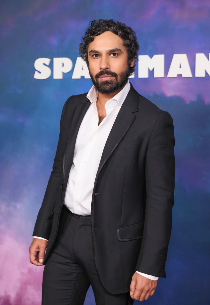 Kunal Nayyar in a black suit and white shirt poses in front of a "Spaceman" sign
