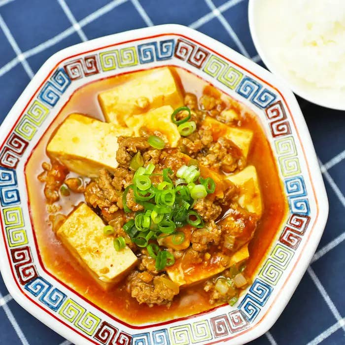 カラフルな皿に盛られたマーボー豆腐が中央にあり、豆腐、ひき肉、ネギが入っている。右上にご飯の皿も見える。