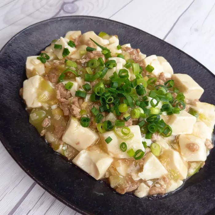 豆腐と野菜、ひき肉を使用した料理が黒い皿に盛り付けられている。上に刻みネギがのっている。