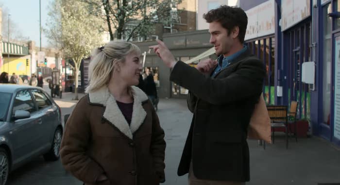 Florence Pugh and Andrew Garfield share a cheerful conversation on a city street. Florence wears a casual jacket, while Andrew is in a blazer and shirt in a scene from We Live in Time