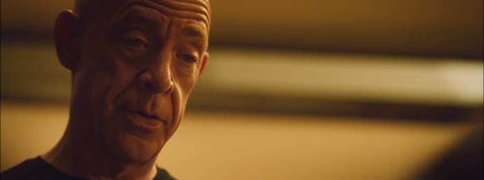 J.K. Simmons is in a close-up shot, wearing a plain shirt, with a neutral expression in an indoor setting
