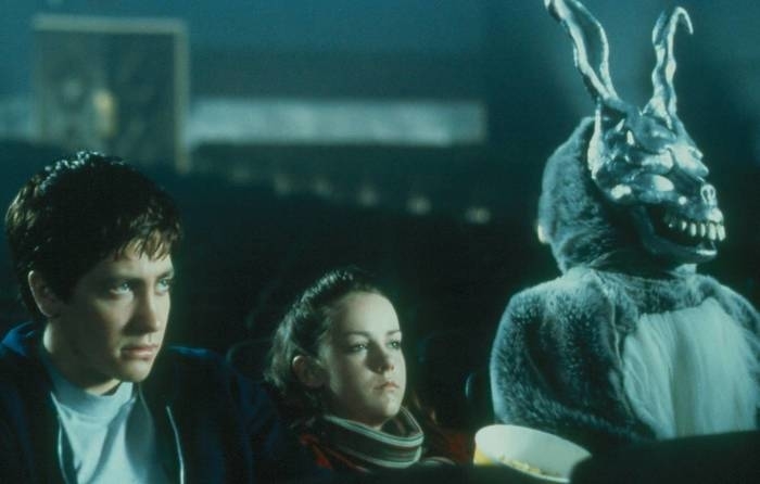 Jake Gyllenhaal, Jena Malone, and a person in a creepy rabbit costume sit in a dark movie theater looking uneasy in a scene from a movie