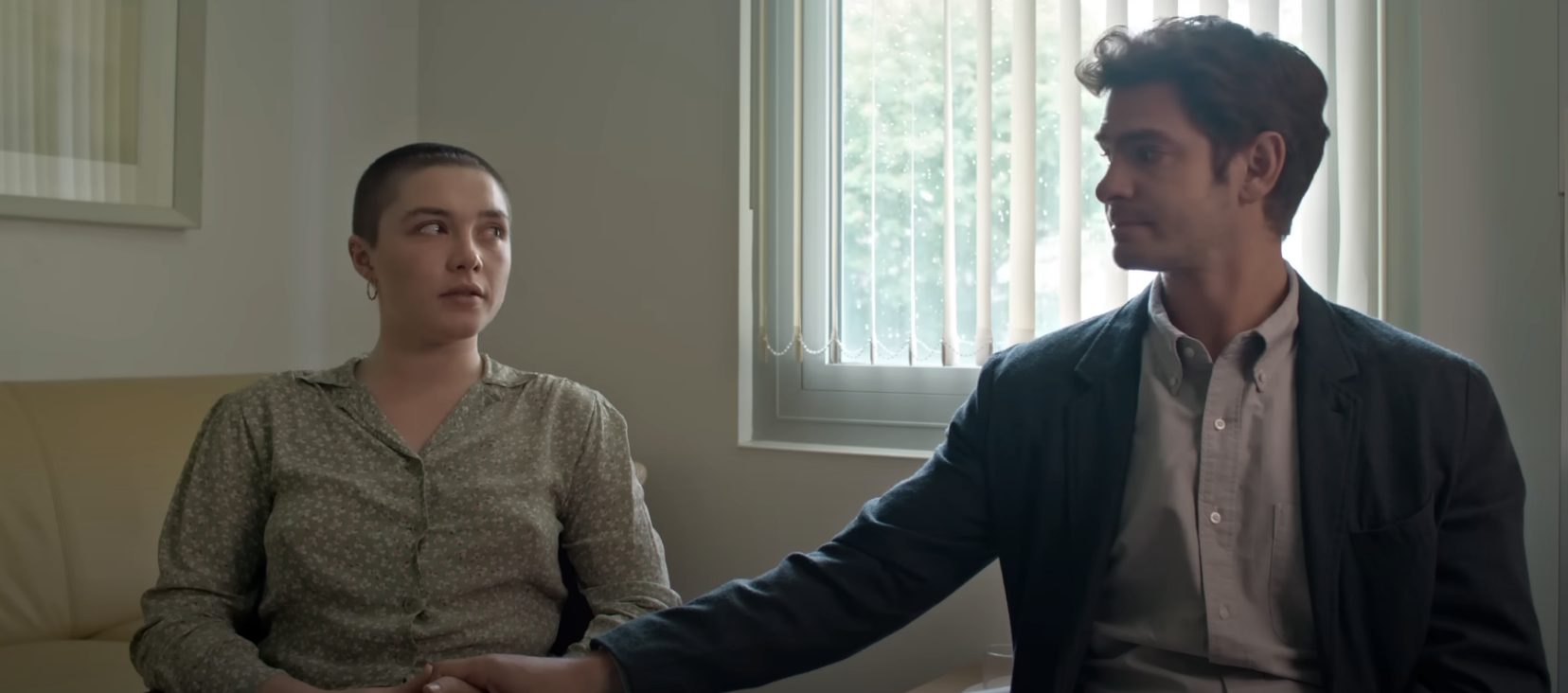 Florence Pugh and Andrew Garfield sit on a couch in a dimly lit room, holding hands, with light streaming through a window behind them