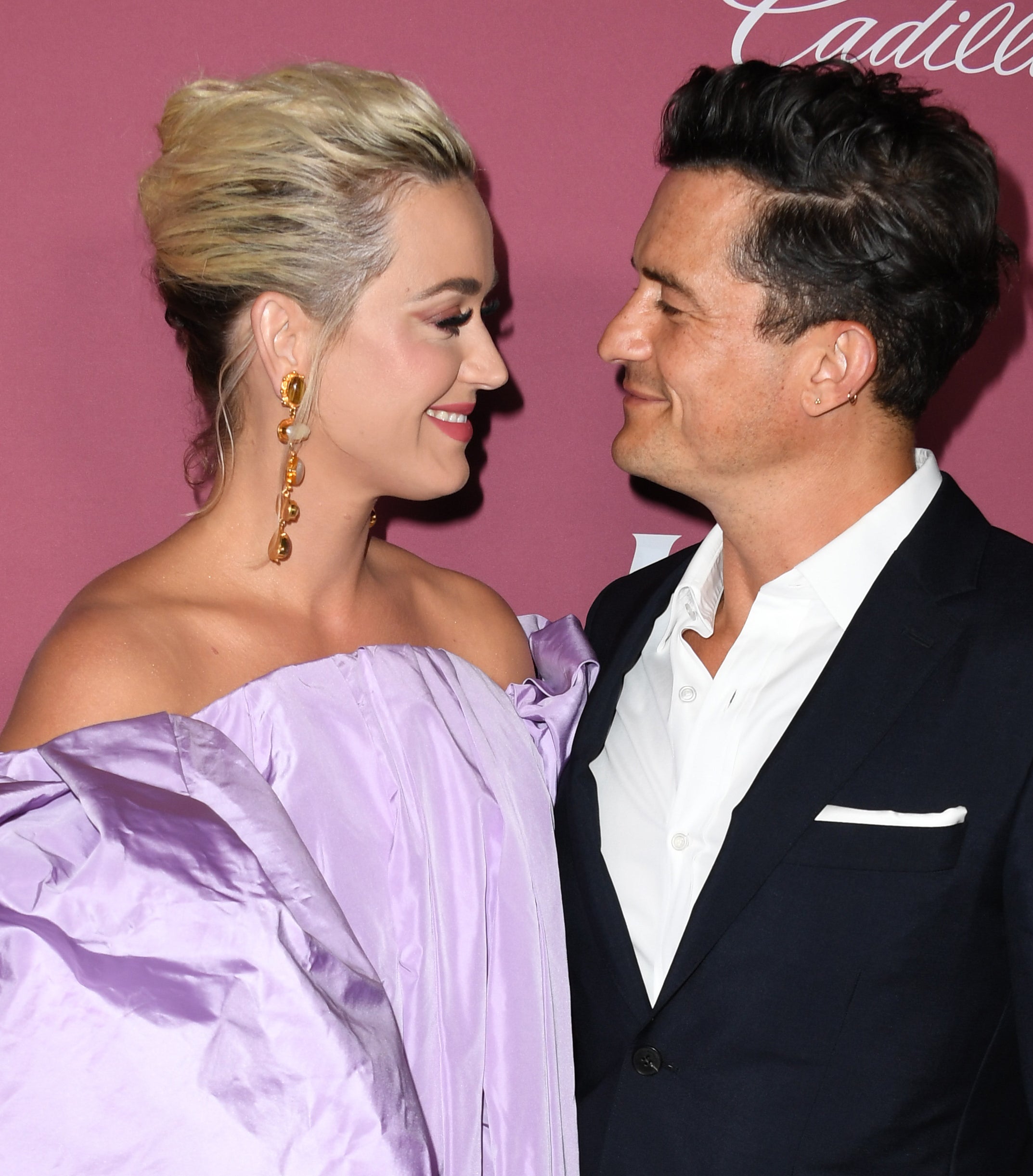 Katy Perry in an off-the-shoulder, billowing gown and Orlando Bloom in a suit, gazing at each other, on a red carpet in front of Cadillac branding