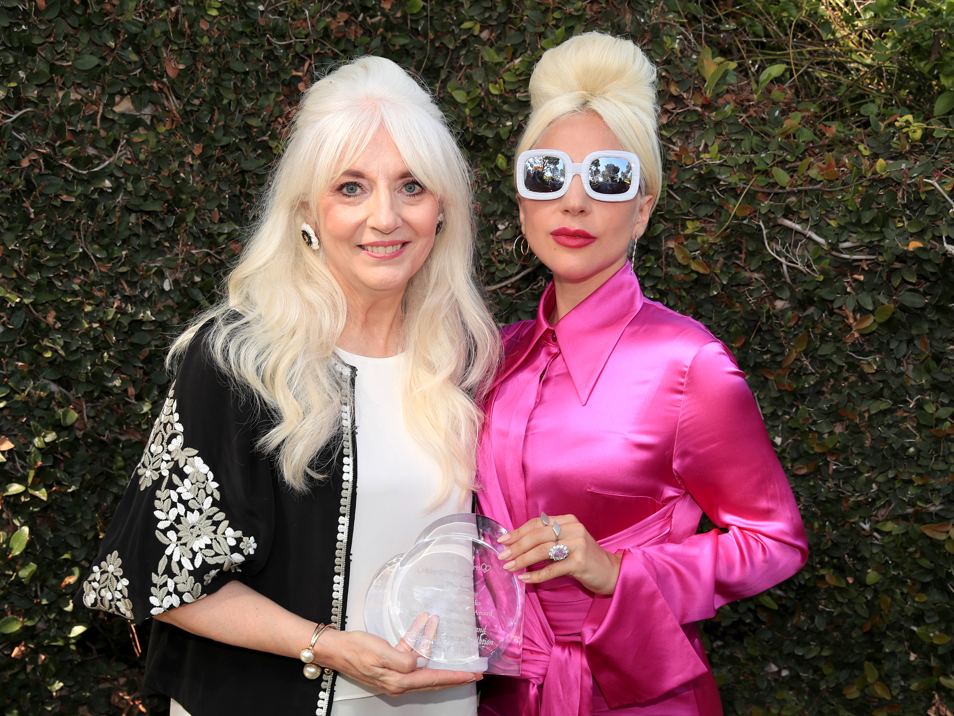 Cynthia Germanotta and Lady Gaga posing together; Cynthia holds an award, and Lady Gaga wears a shiny outfit with large sunglasses