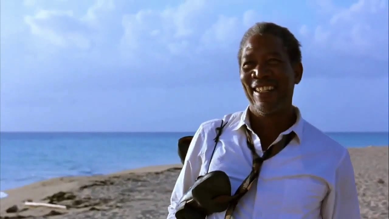 Morgan Freeman smiles on a sandy beach, wearing a white shirt and a loosened black tie draped over his shoulder