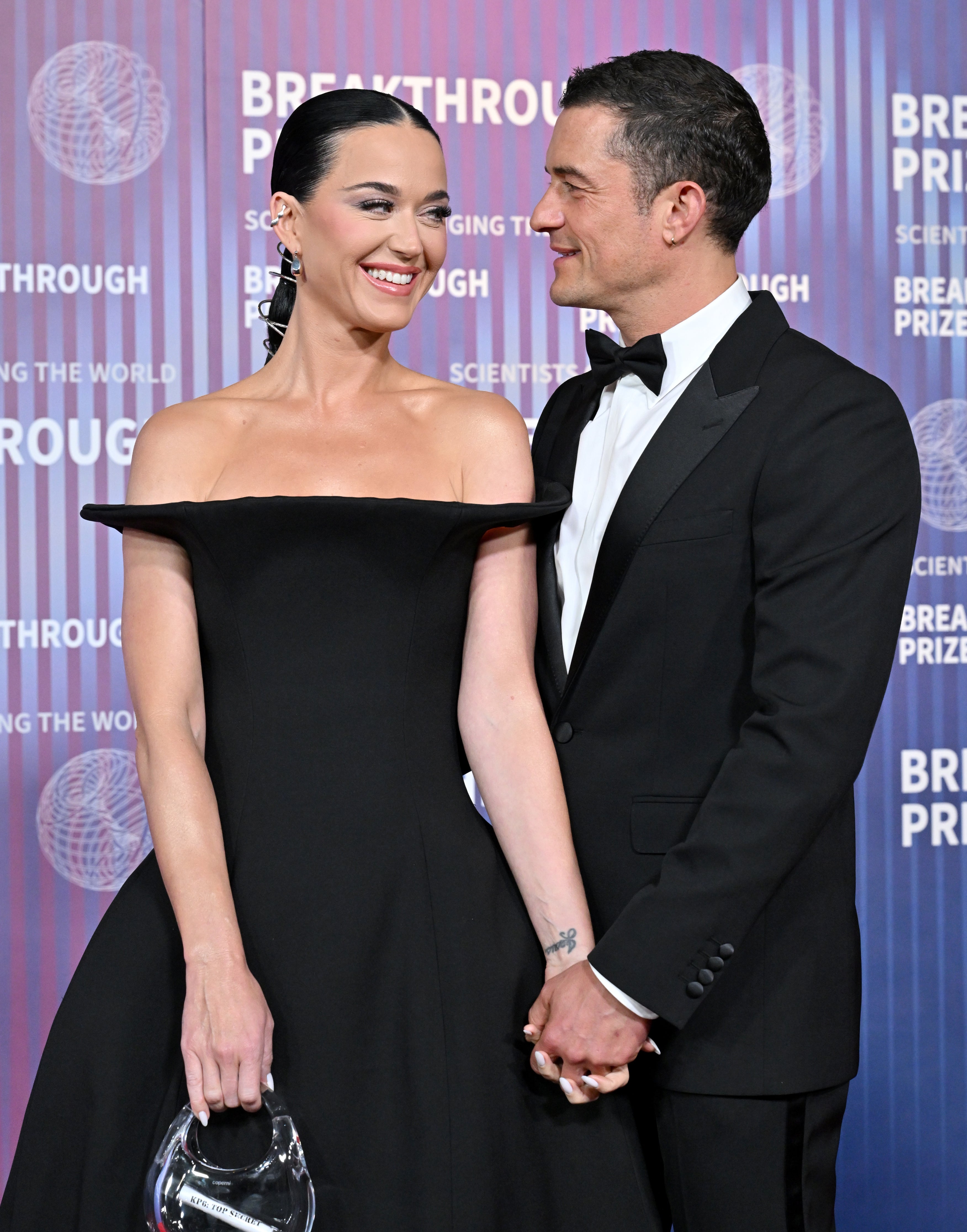 Katy Perry and Orlando Bloom smiling at each other and holding hands at the Breakthrough Prize event. Katy is wearing an off-shoulder black gown, and Orlando is in a tuxedo