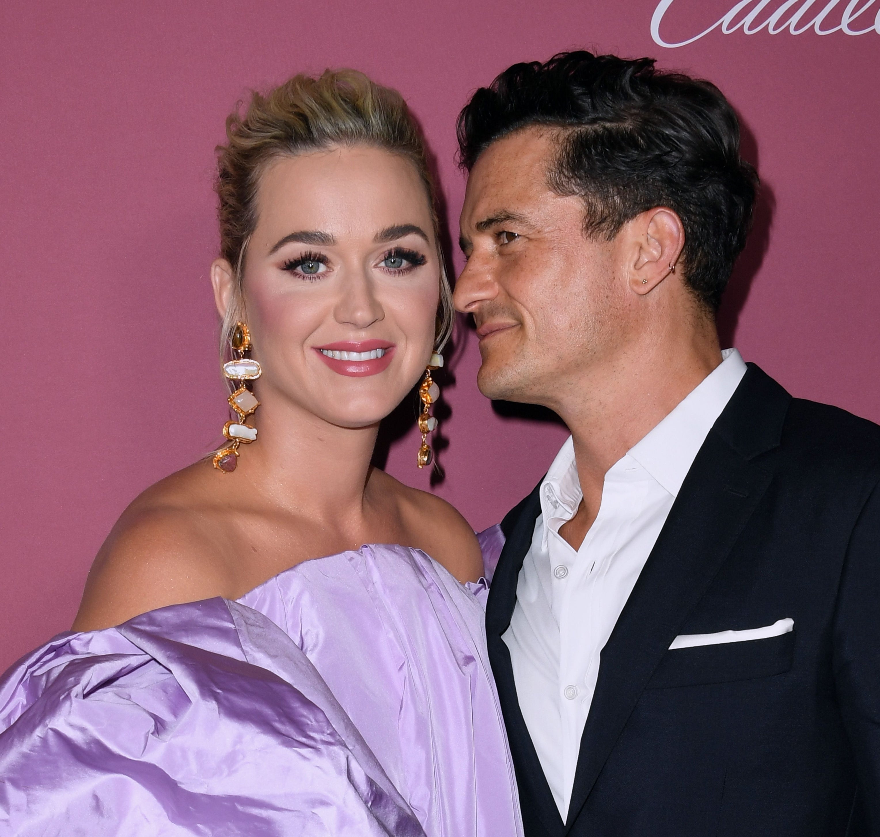 Katy Perry and Orlando Bloom smile on a red carpet. Katy wears a glamorous off-shoulder dress with statement earrings. Orlando is in a black suit