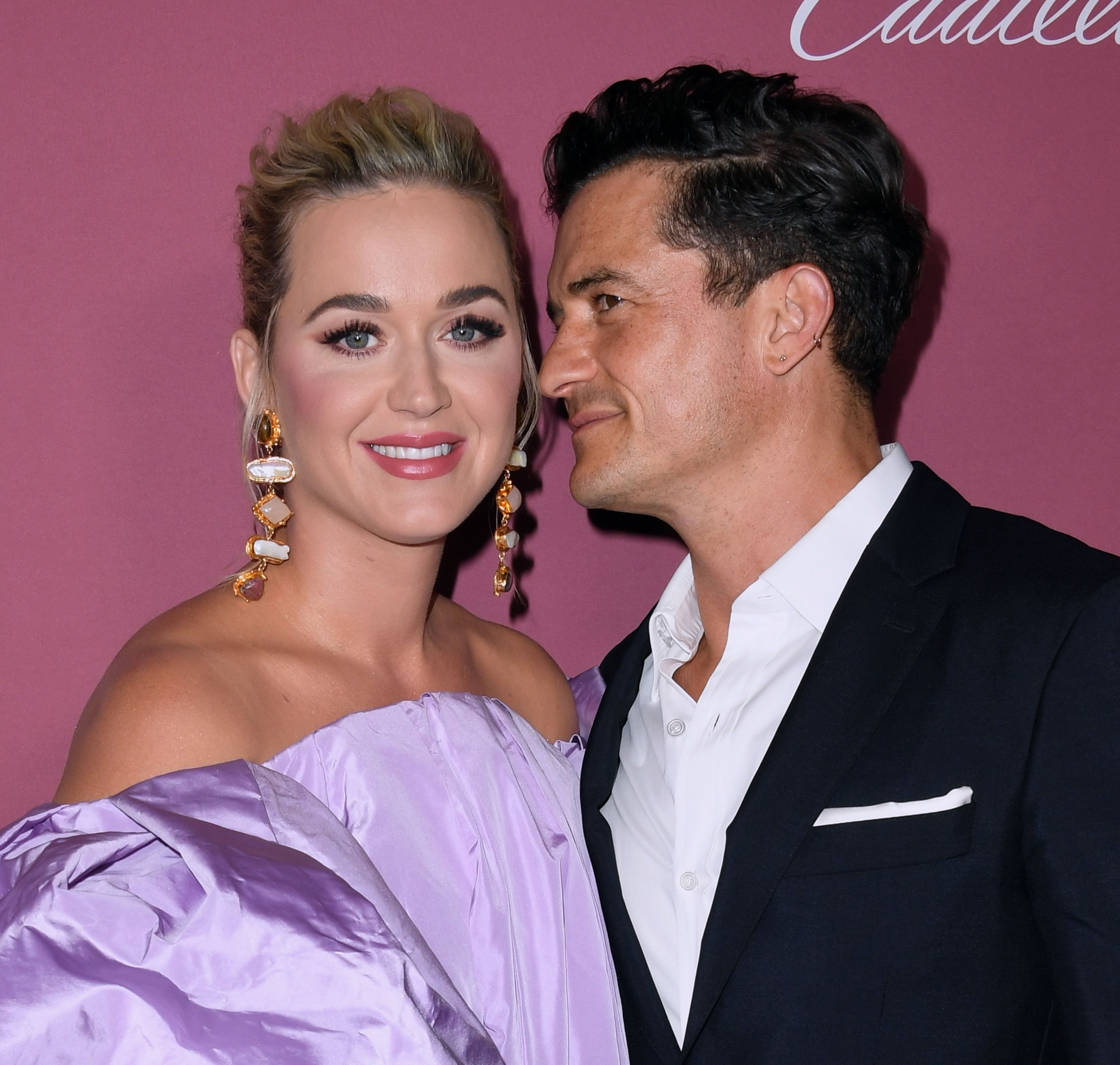 Katy Perry and Orlando Bloom smile on a red carpet. Katy wears a glamorous off-shoulder dress with statement earrings. Orlando is in a black suit