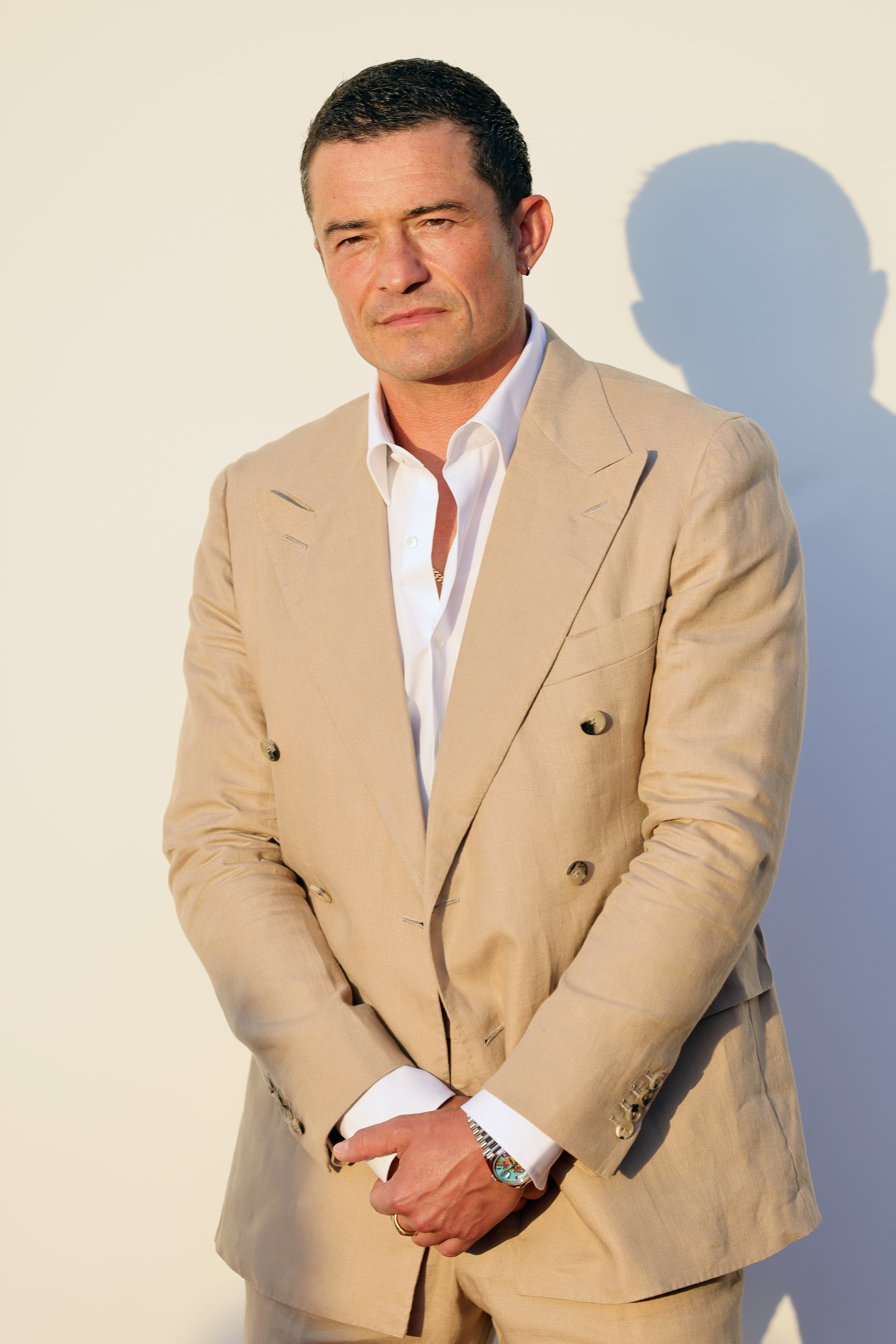 Orlando Bloom in a light suit with a white shirt, standing against a plain background, hands clasped