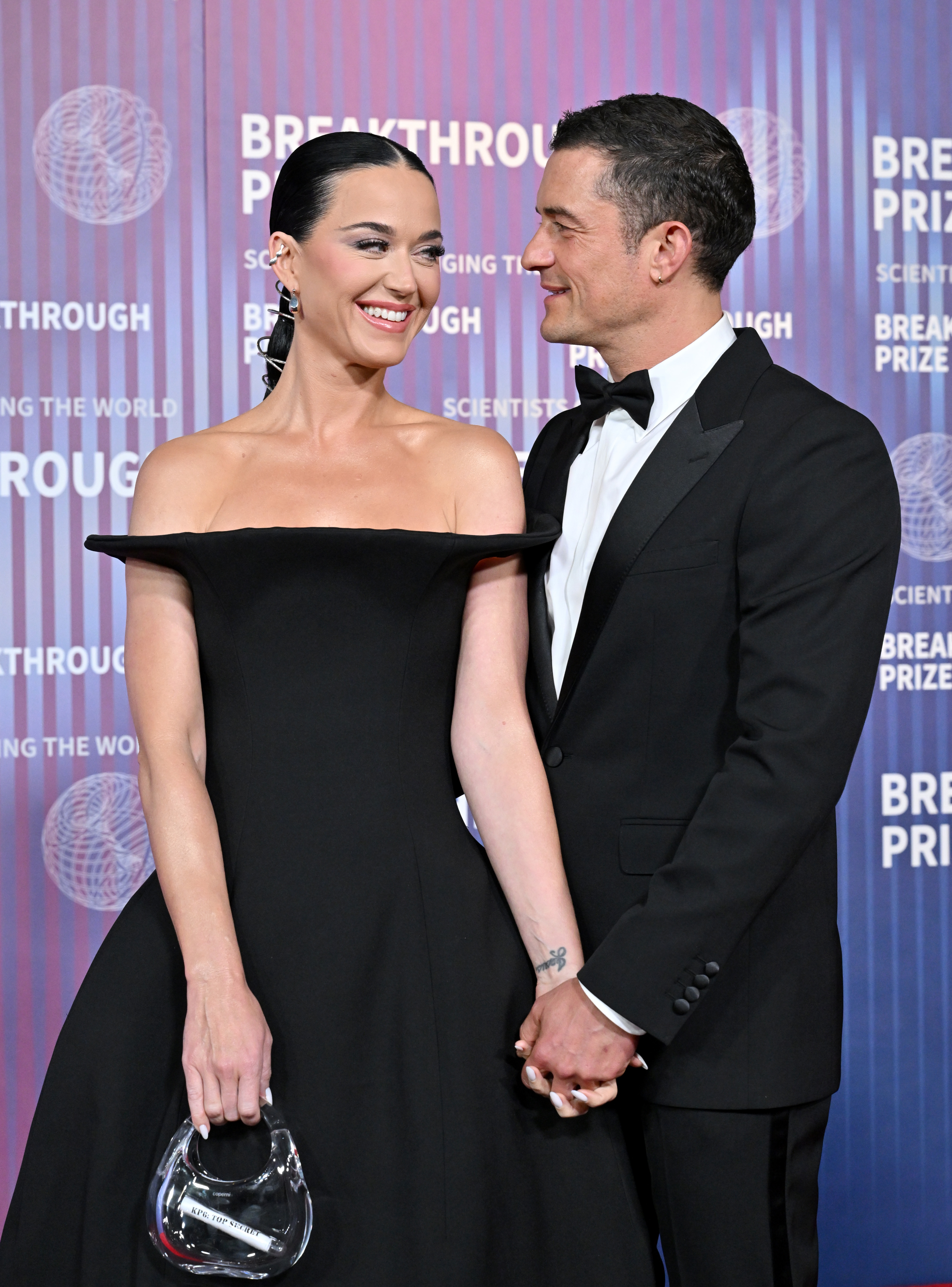 Katy Perry and Orlando Bloom smiling and holding hands on a red carpet at the Breakthrough Prize event. Katy is wearing an off-shoulder black gown