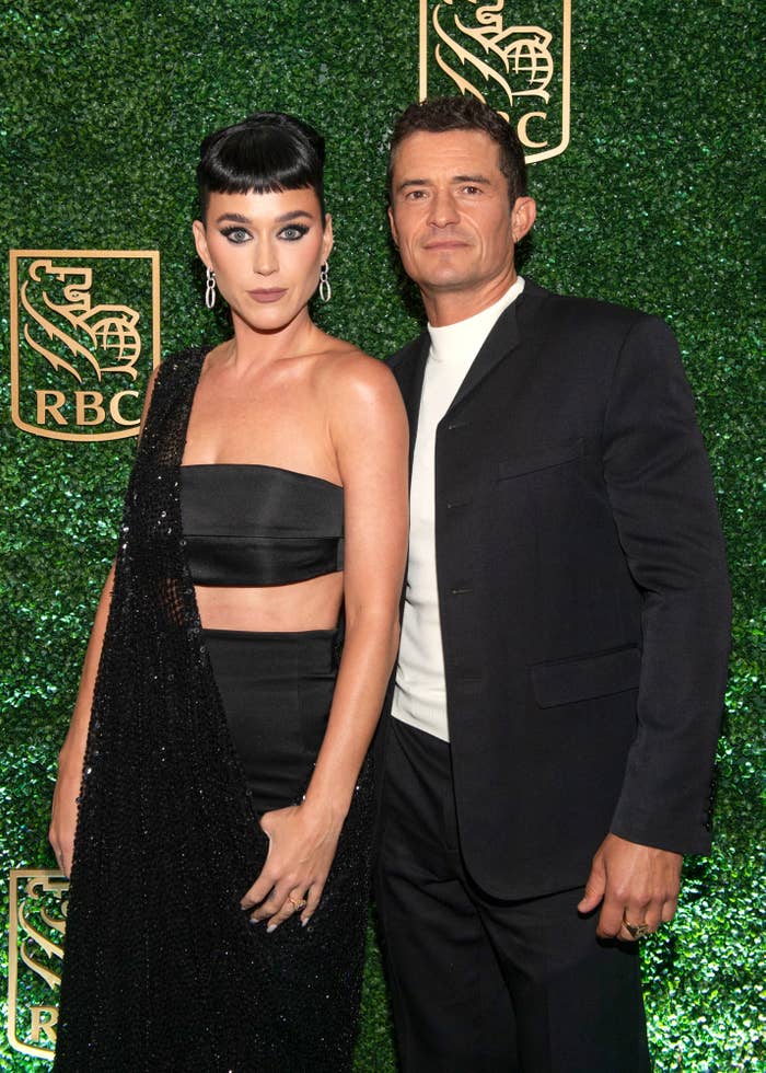 Katy Perry and Orlando Bloom pose together, with Katy wearing a black crop top and long skirt, and Orlando in a black suit with a white shirt, against a green backdrop