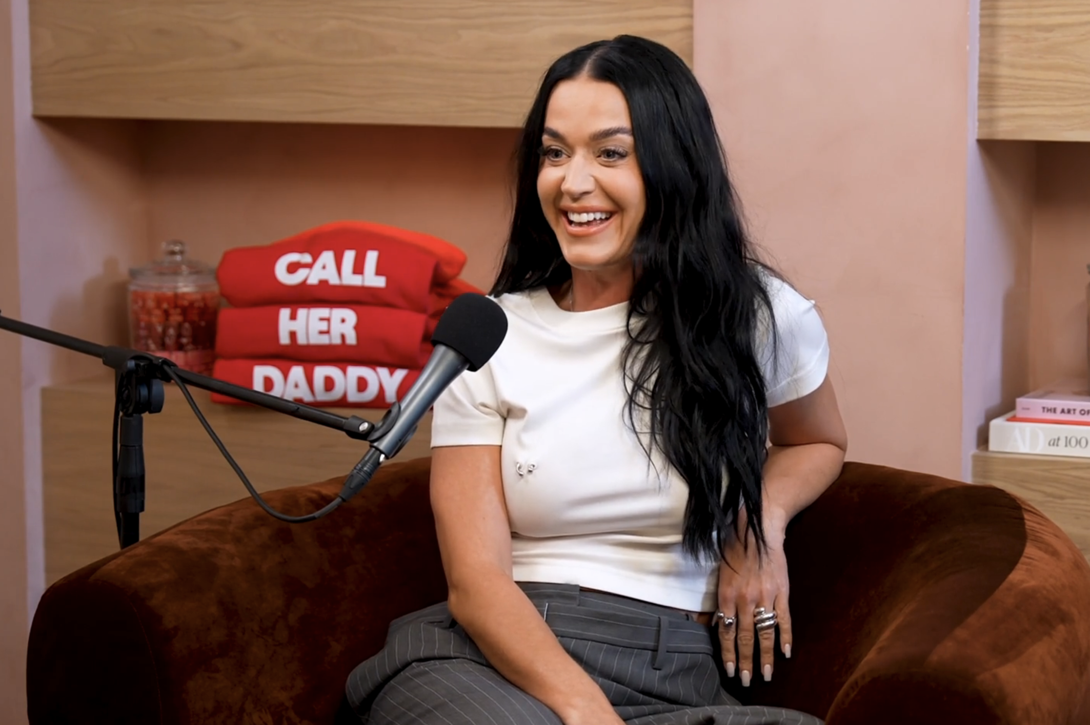 Katy Perry in a white top and pinstripe pants, smiling during a podcast interview with a &quot;Call Her Daddy&quot; backdrop