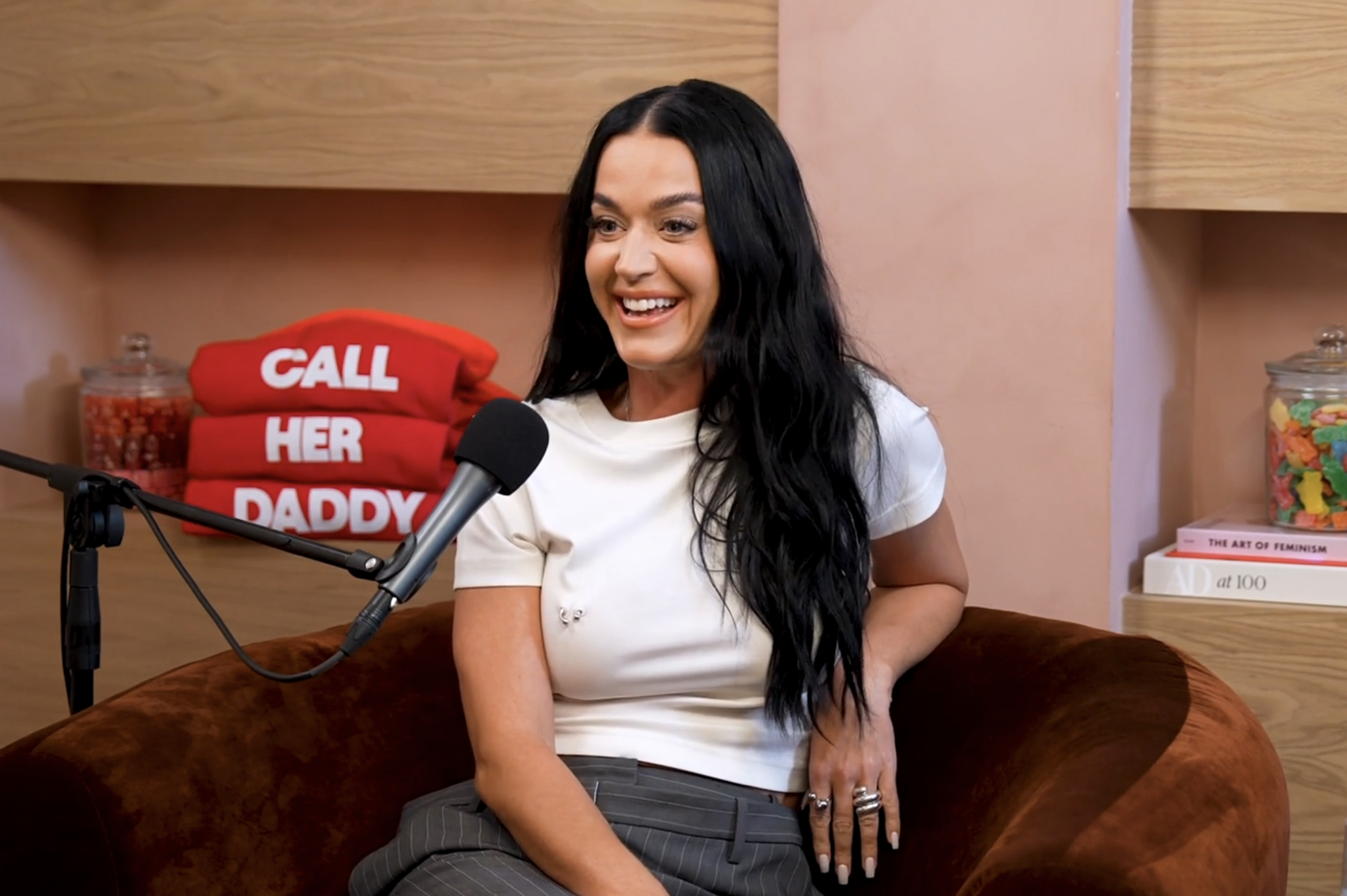 Katy Perry in a white top and pinstripe pants, smiling during a podcast interview with a "Call Her Daddy" backdrop