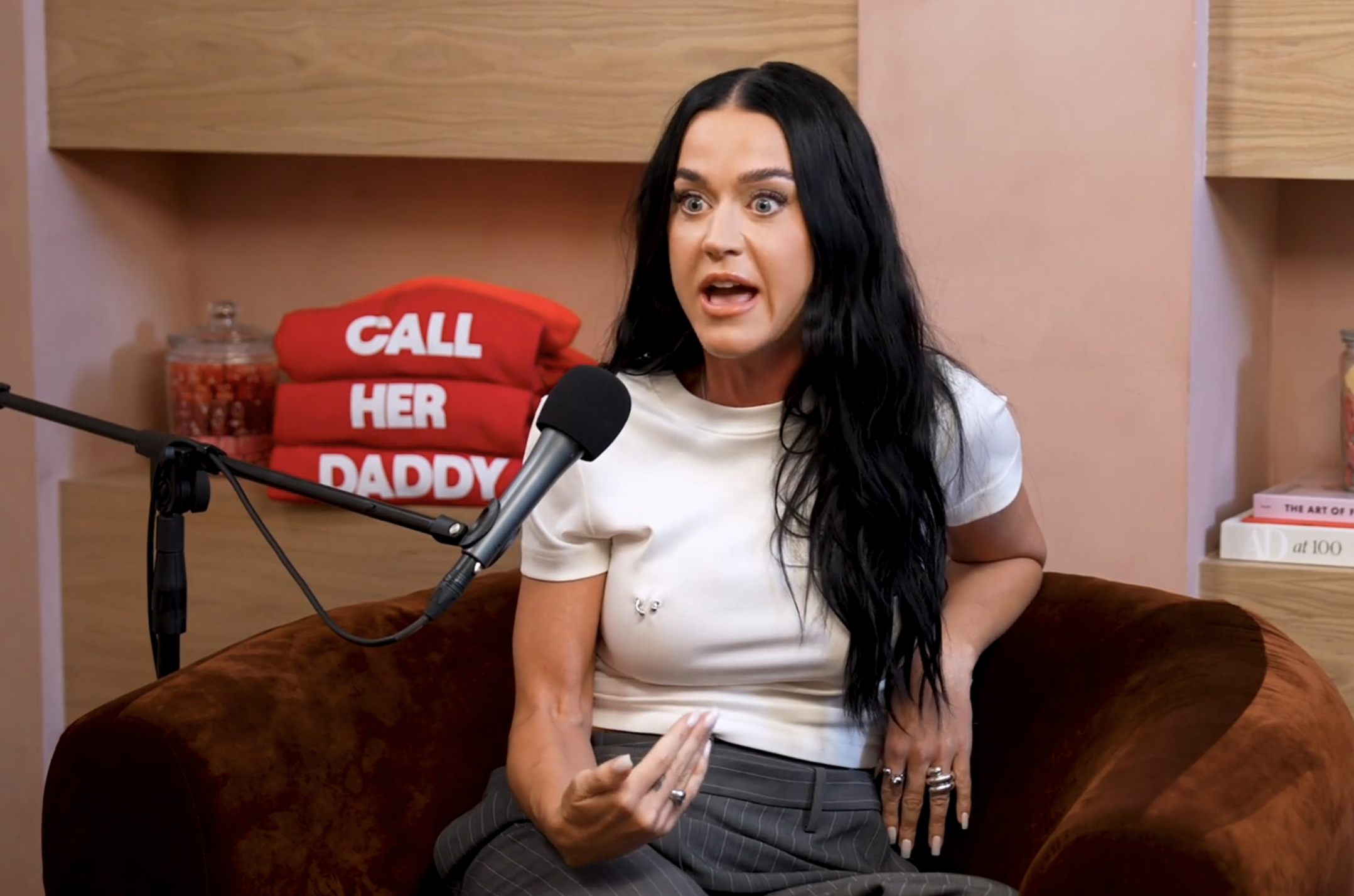 Katy Perry on the &quot;Call Her Daddy&quot; podcast, speaking into a microphone while seated on a brown chair. Shelves with candy jars and books are in the background