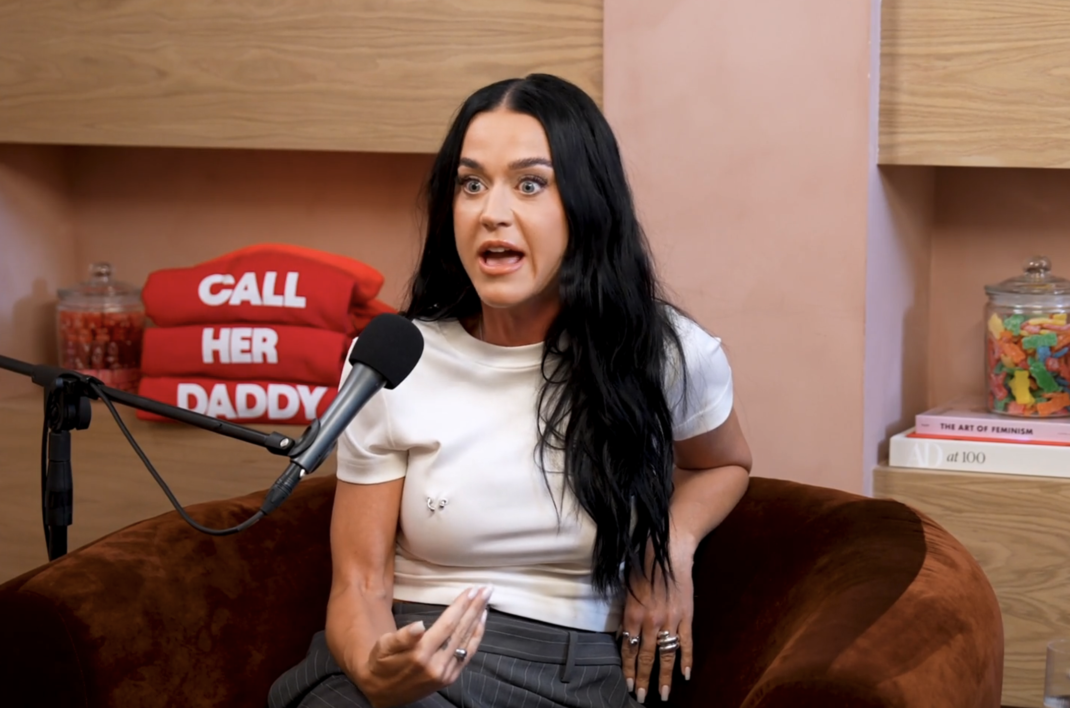 Katy Perry on the "Call Her Daddy" podcast, speaking into a microphone while seated on a brown chair. Shelves with candy jars and books are in the background