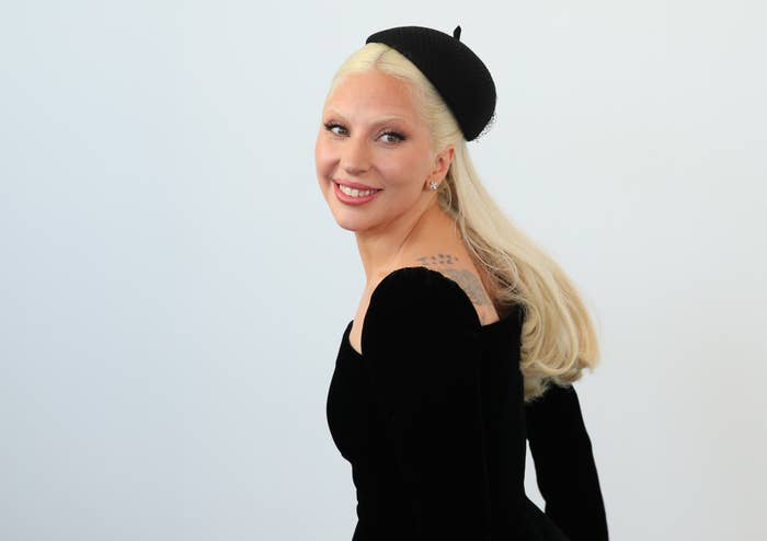 Lady Gaga smiling, facing slightly to her left, wearing a black dress and a matching black hat. Her blonde hair is worn down