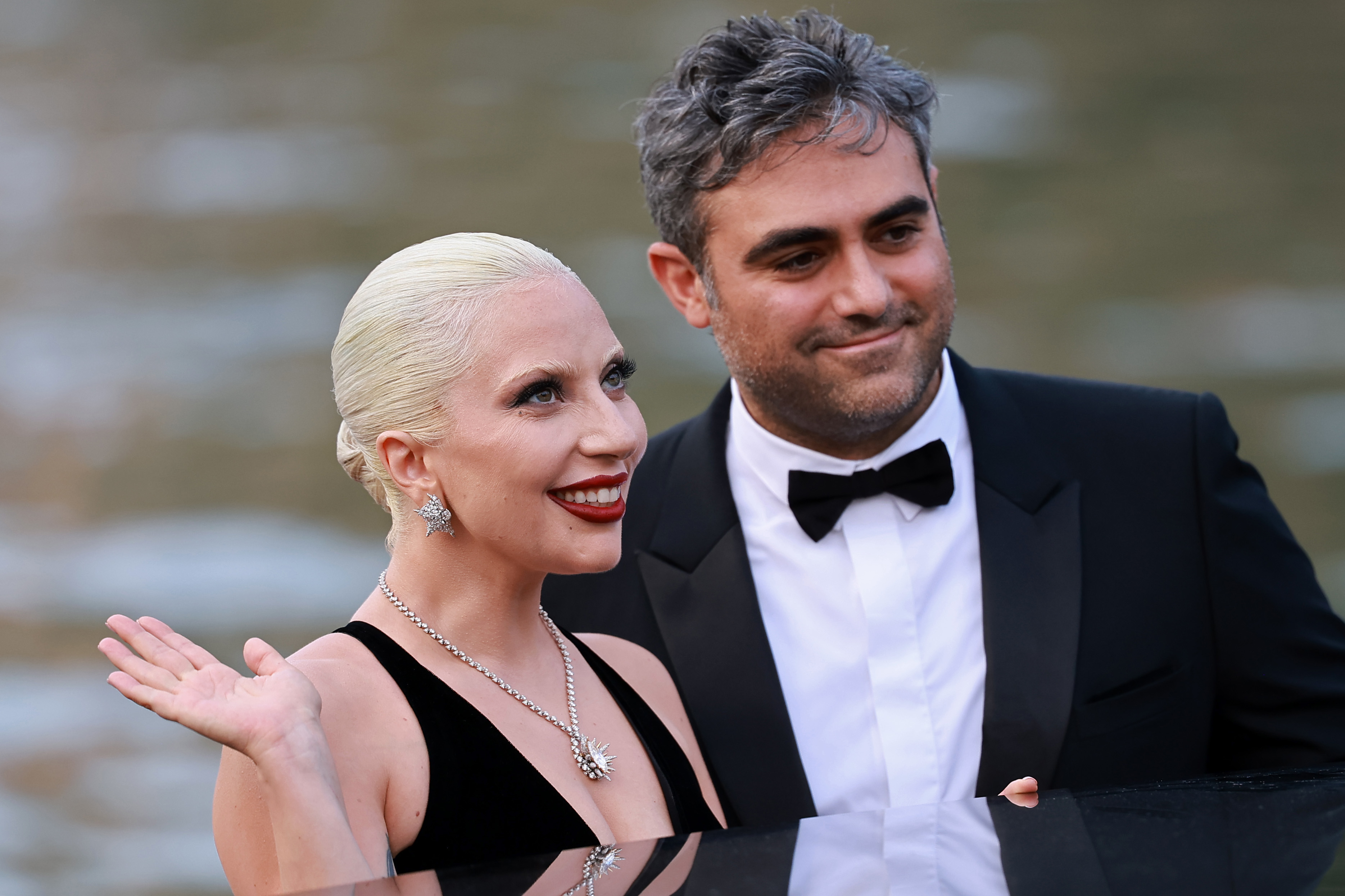 Lady Gaga, in an elegant black dress and diamond jewelry, waves while standing next to Michael Polansky in a tuxedo by a body of water