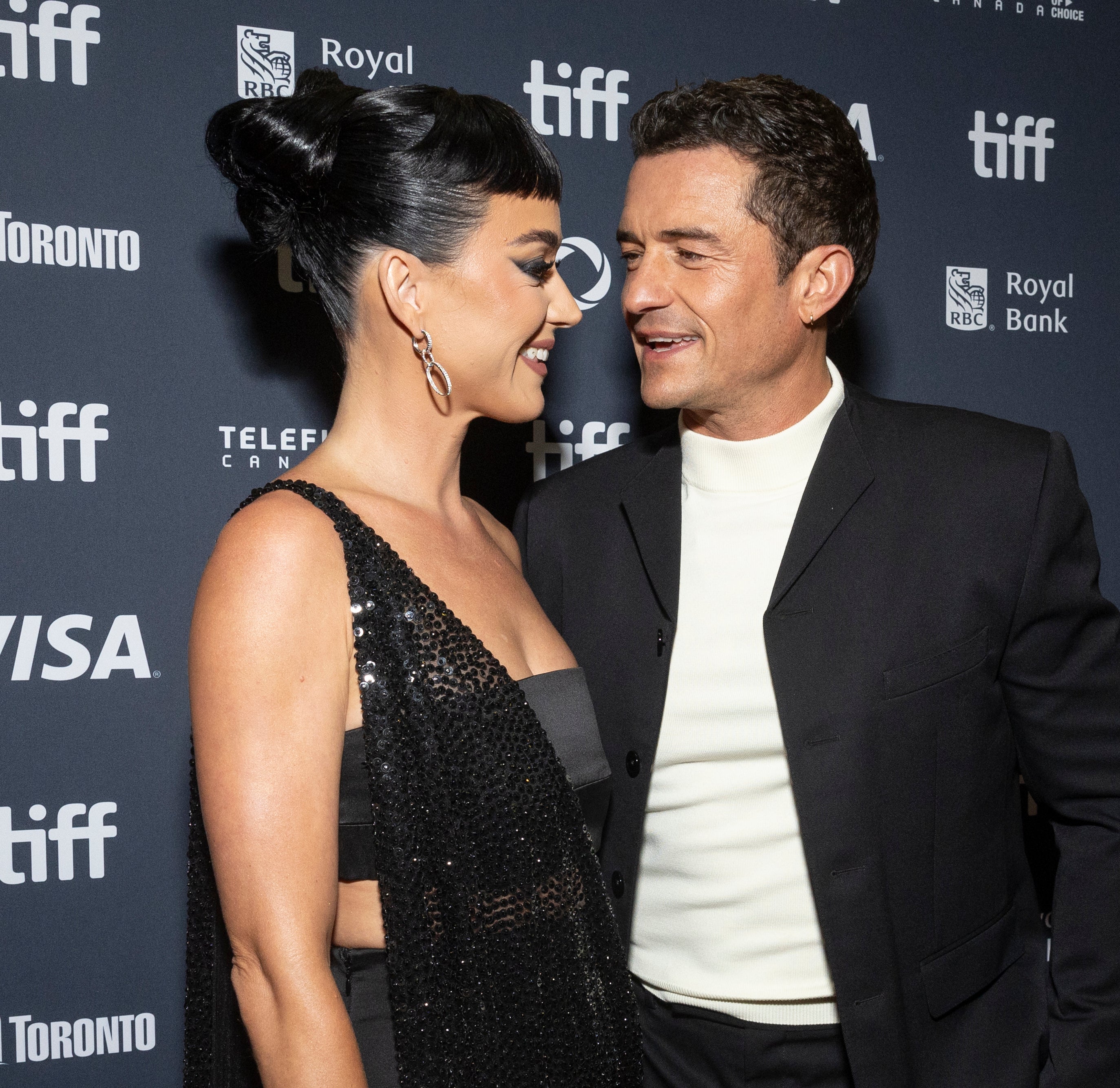 Katy Perry in a dazzling black gown and Orlando Bloom in a black suit with a white shirt smile at each other on the TIFF red carpet