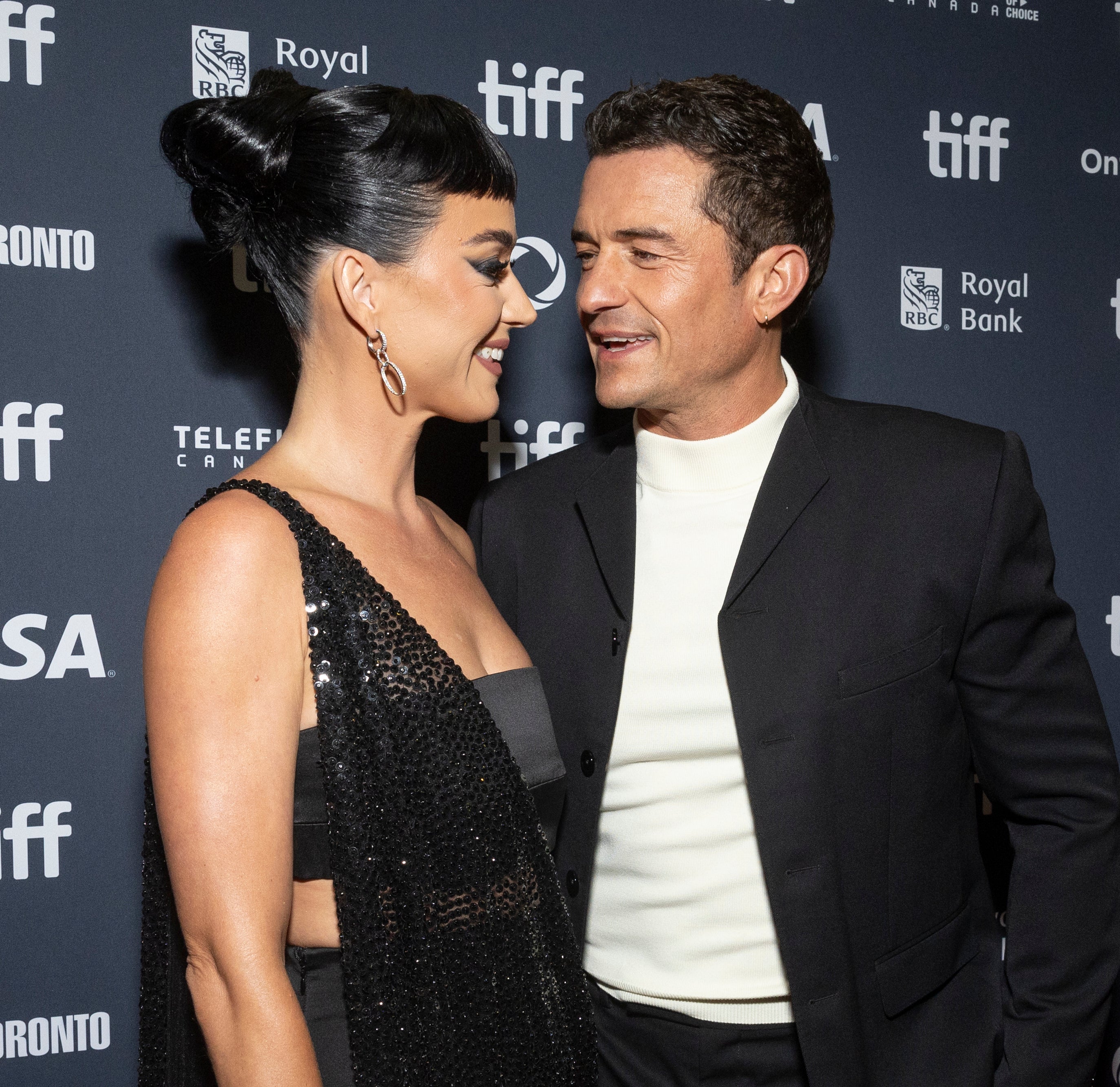 Katy Perry in a dazzling black gown and Orlando Bloom in a black suit with a white shirt smile at each other on the TIFF red carpet