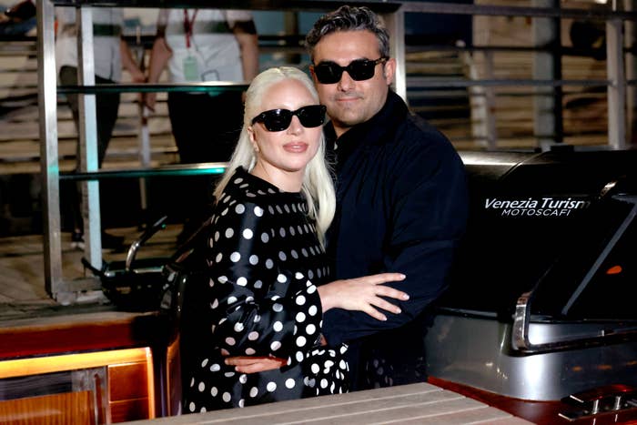 Lady Gaga in a polka-dot dress poses with Michael Polansky, both wearing dark sunglasses, near a boat docked at Venezia Turisms Motoscafi
