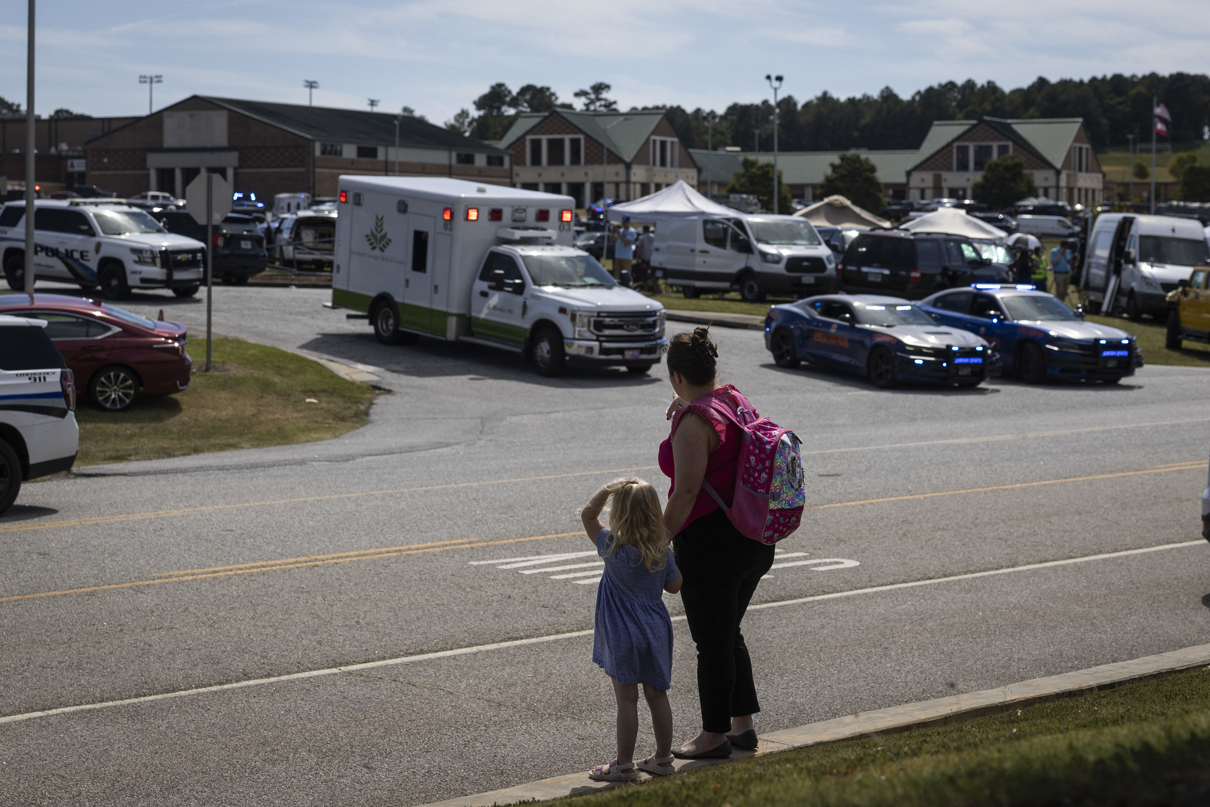 A woman with a backpack and a child stand near emergency vehicles, including an ambulance and police cars, outside a school-like building