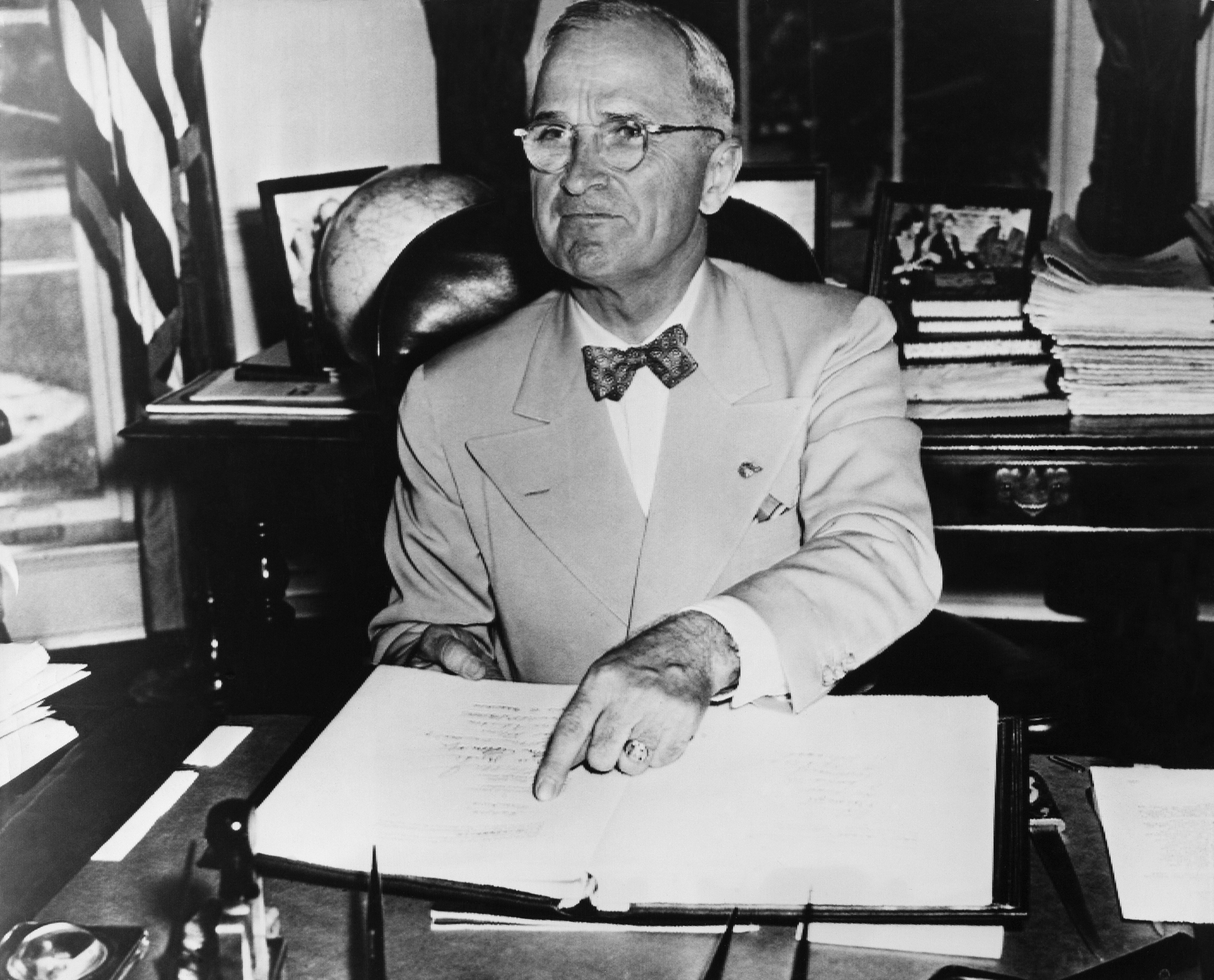 Harry S. Truman, dressed in a suit, sits at a desk, pointing to an open book, surrounded by papers and photos in an office