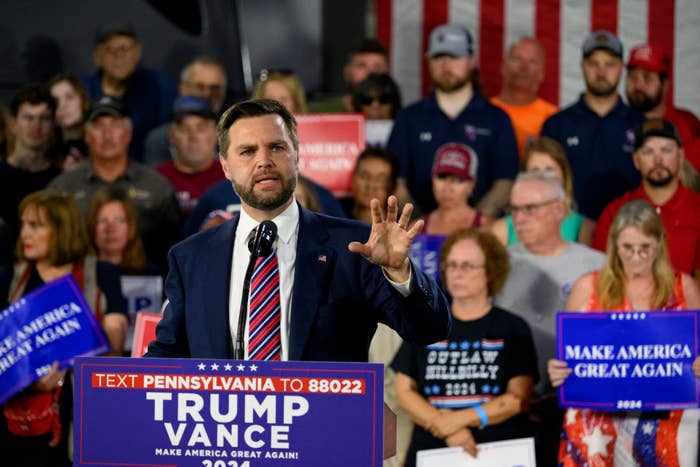 J.D. Vance speaks at a podium during a rally with people holding &quot;Make America Great Again&quot; signs and a US flag backdrop