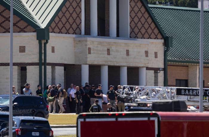 Group of people, including law enforcement, gather outside a building with a green metal roof, amid emergency response vehicles in the foreground
