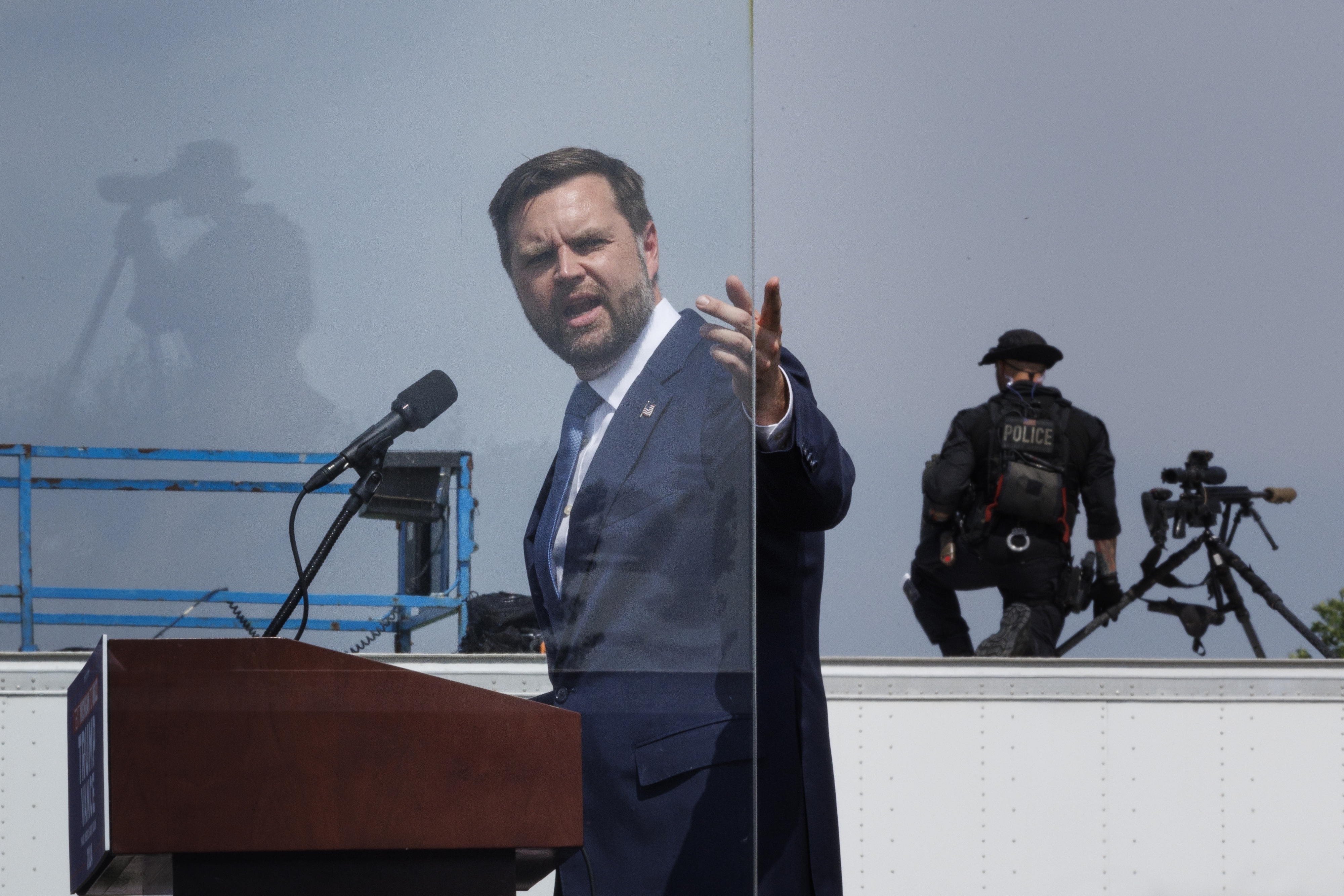 J.D. Vance speaks at a podium behind a transparent barrier, while police officers and media equipment are visible in the background