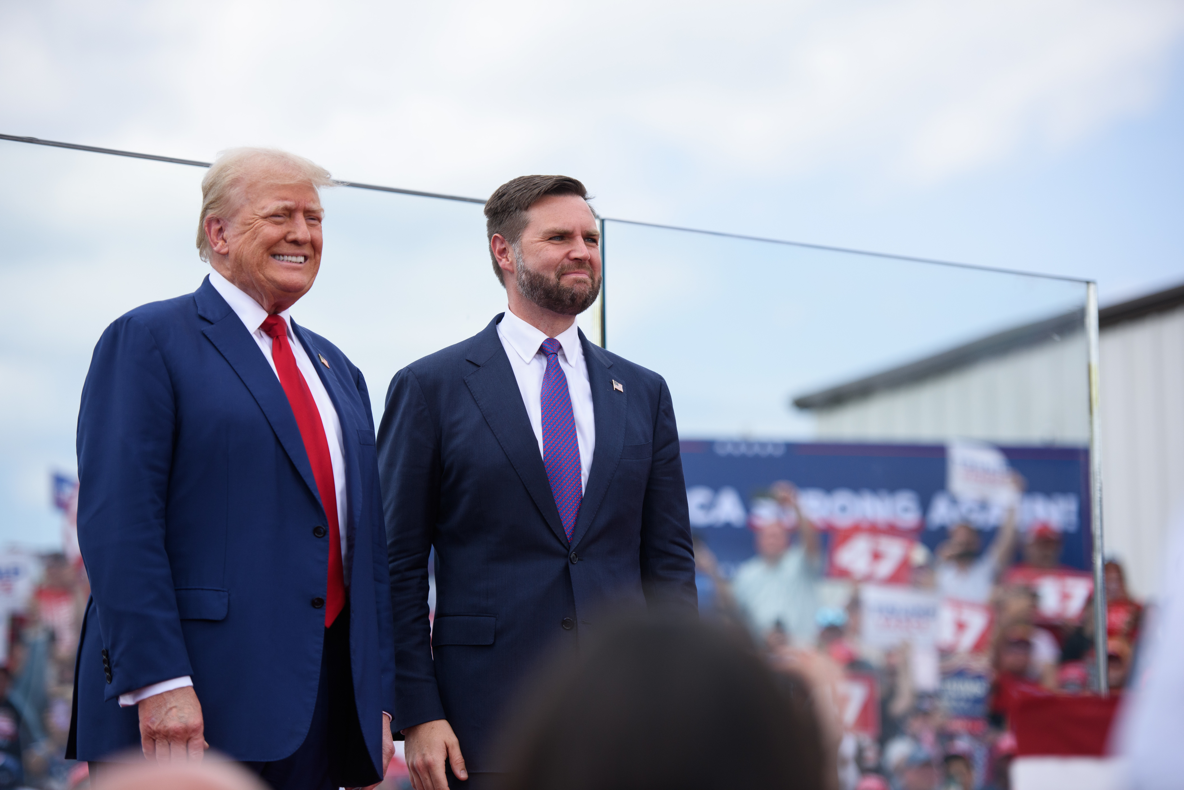 Donald Trump and J.D. Vance at a rally, standing side by side, both wearing suits and ties. Crowd and signs visible in the background