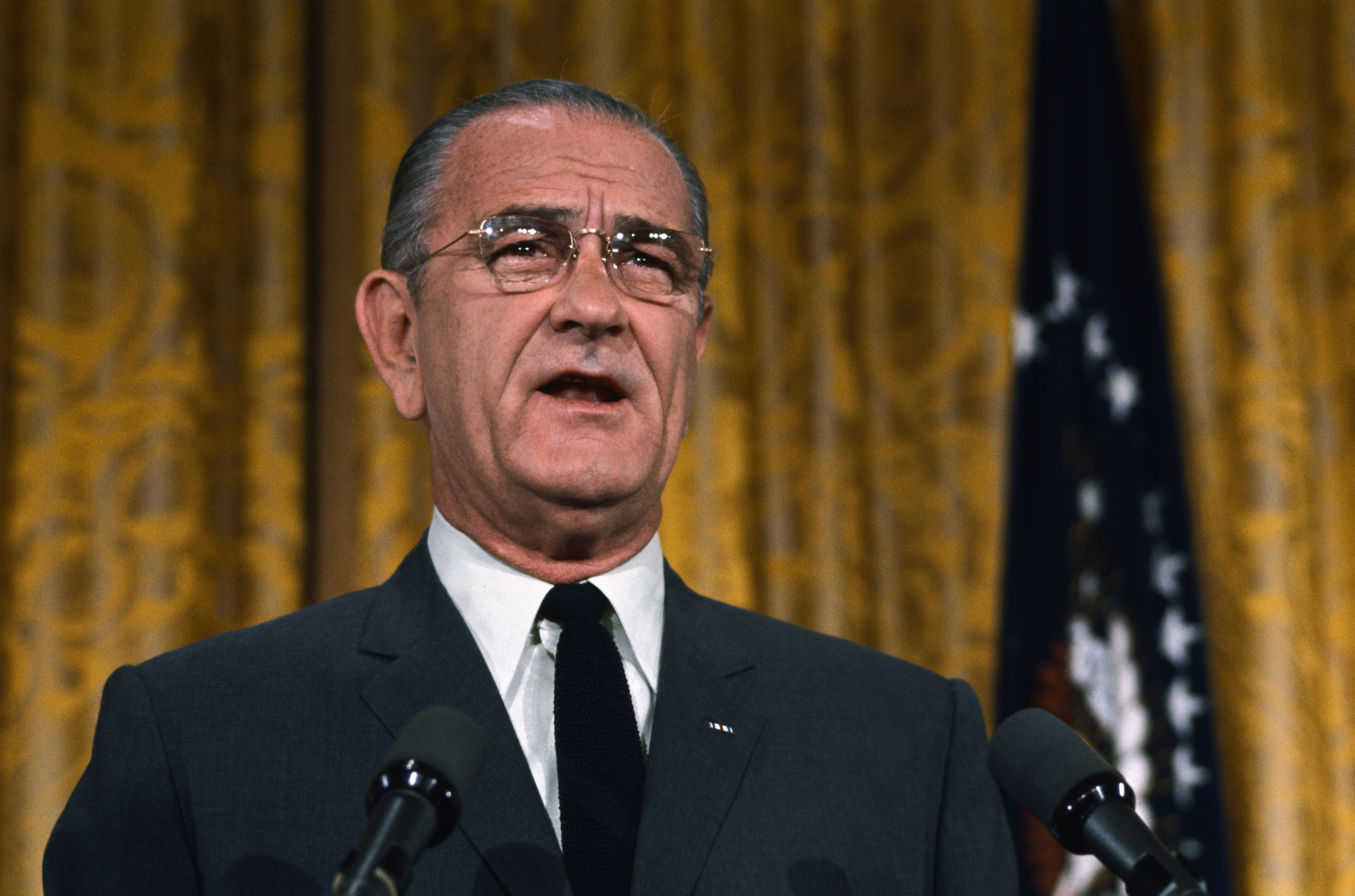 Lyndon B. Johnson speaking at a podium with microphones, dressed in a suit and tie. He is in a room with a patterned curtain backdrop