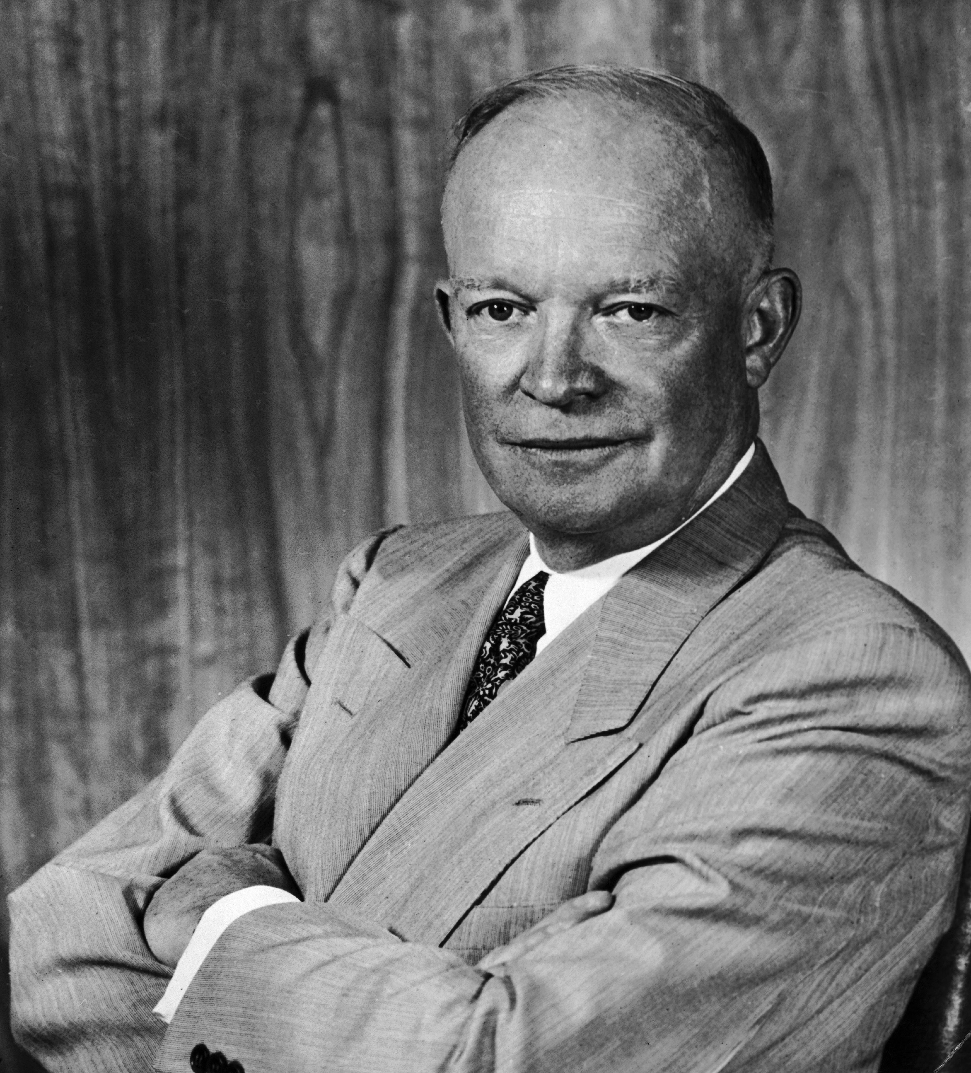 Dwight D. Eisenhower, in a suit with arms crossed, poses for a formal portrait against a plain background