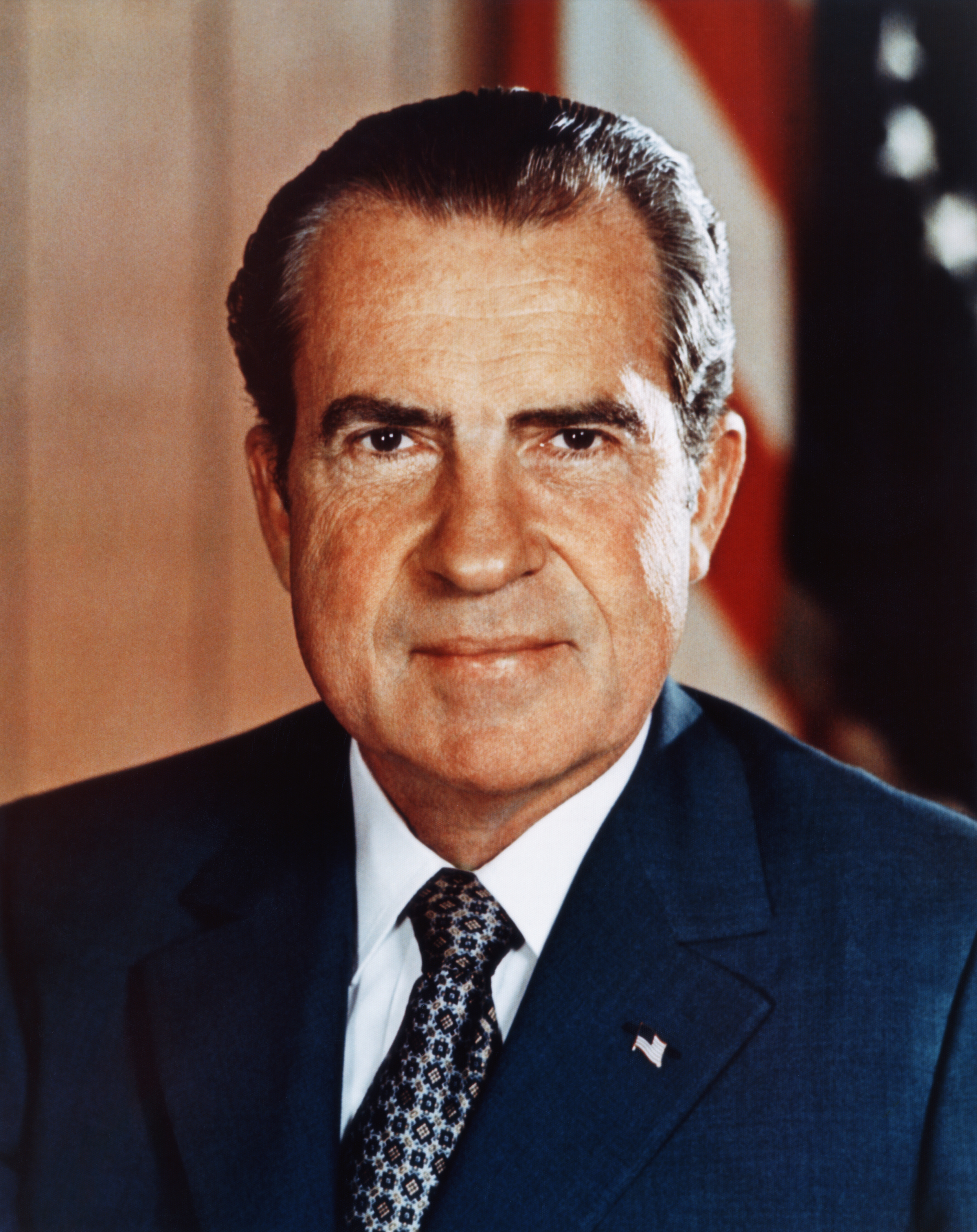 Richard Nixon, wearing a suit and tie, is posed looking directly at the camera with an American flag in the background