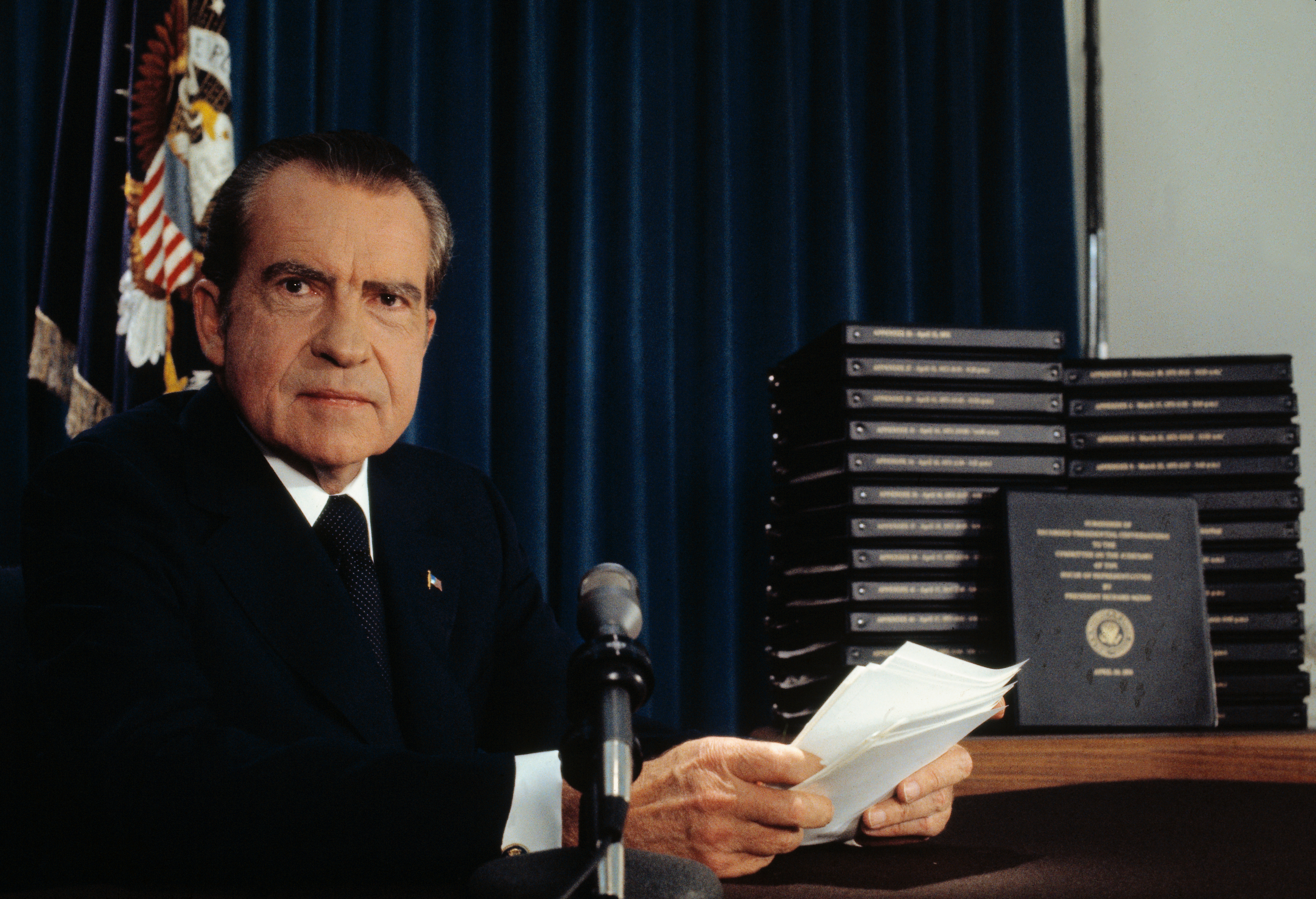 Richard Nixon at a desk holding papers with a stack of binders labeled "Watergate" behind him, flag and presidential seal in the background