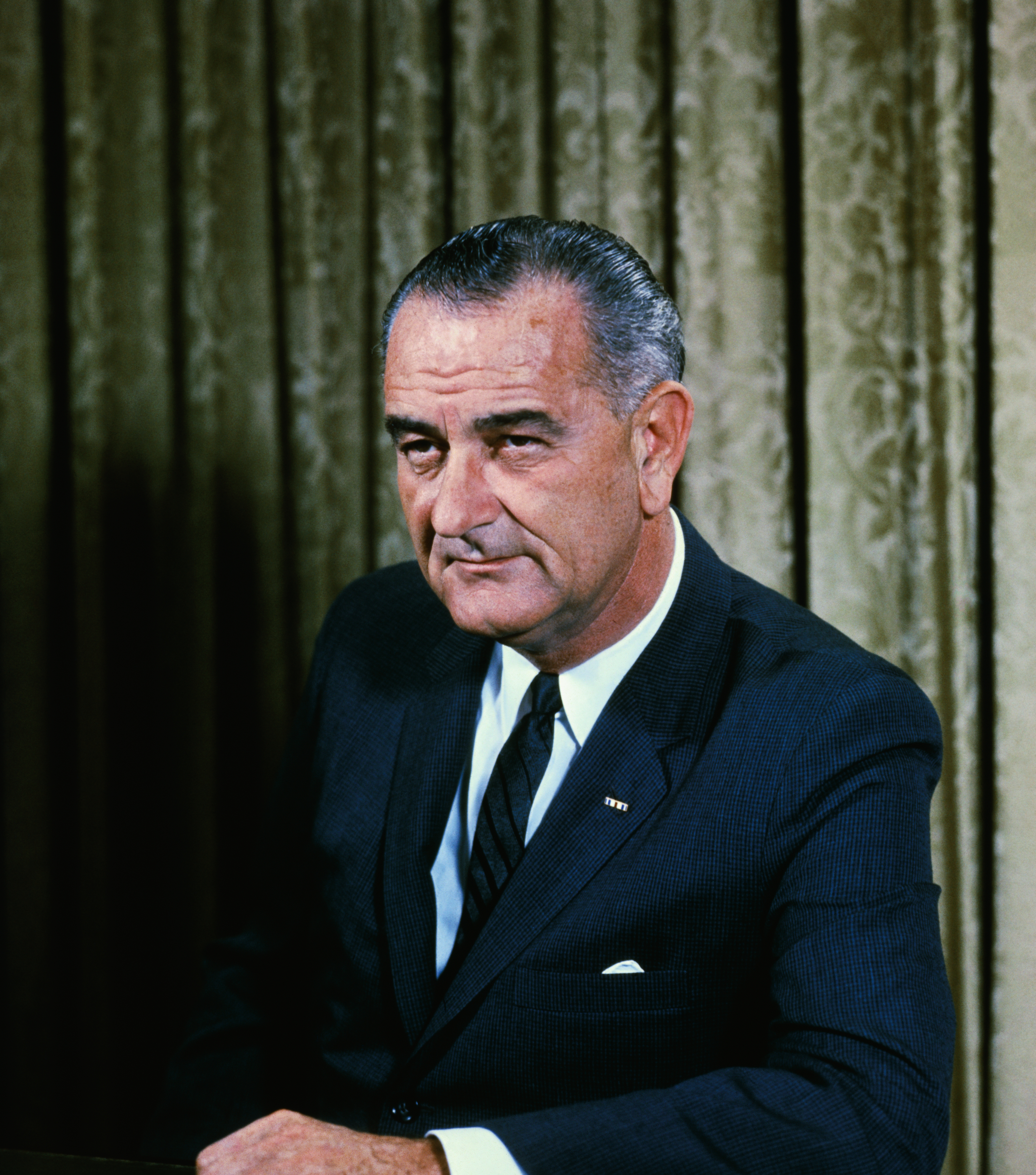 A man in a suit and tie sits at a wooden table in front of a patterned curtain