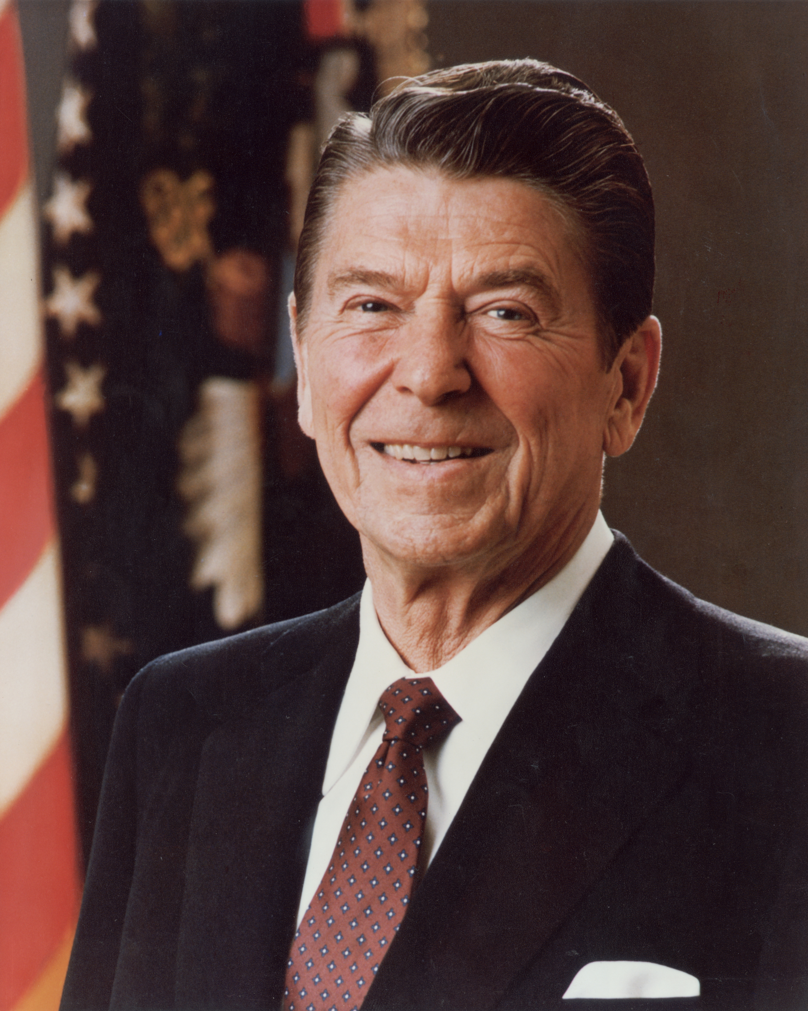 Ronald Reagan, wearing a suit and tie, smiles for an official portrait with a flag in the background